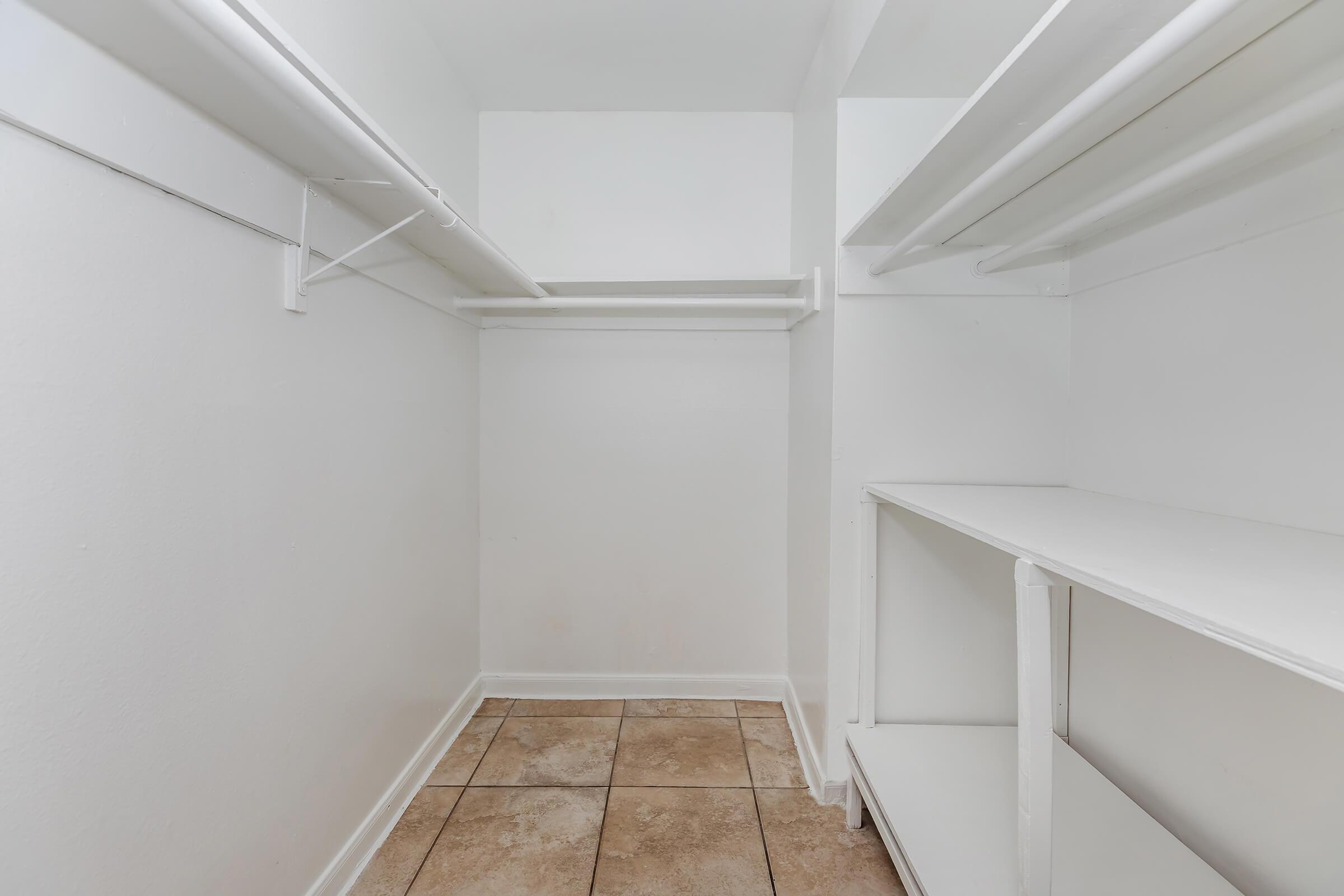 A clean, empty walk-in closet featuring white walls, a tiled floor, and several shelves. The space has hanging rods on one side and built-in shelves on the other, providing ample storage options. The overall appearance is bright and organized, with no clutter.