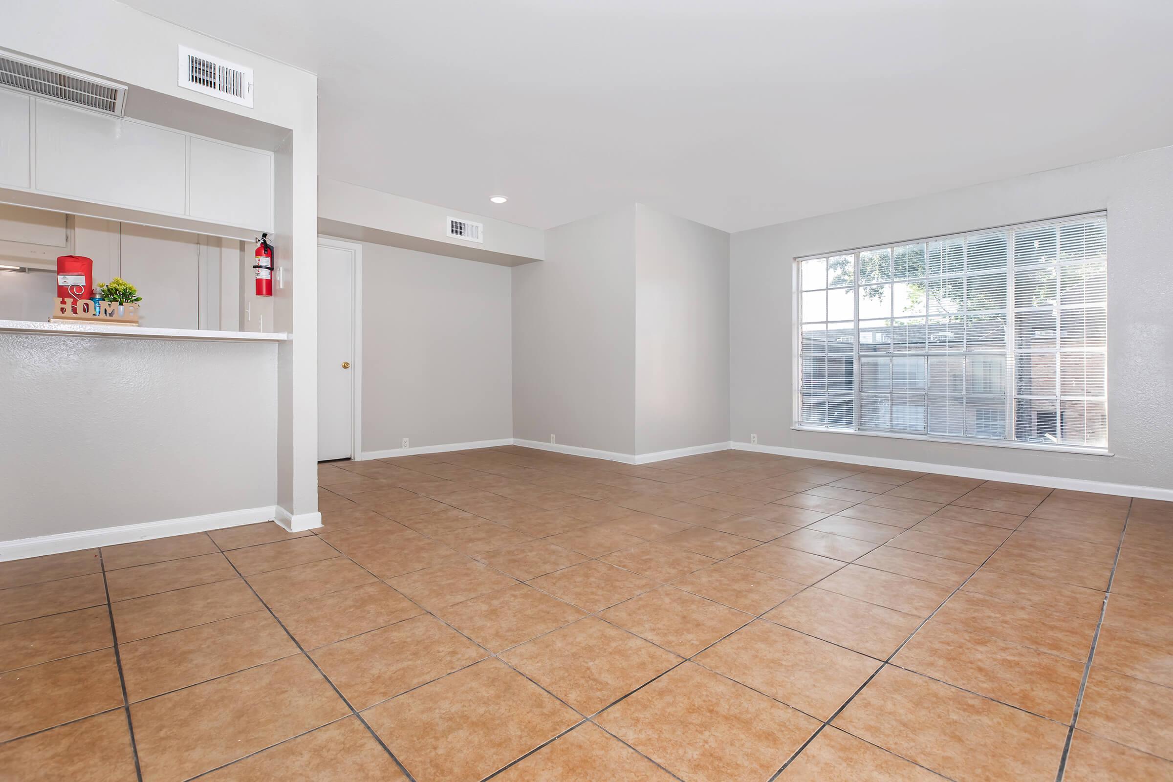 Spacious interior of a modern living room featuring tiled flooring, a large window allowing natural light, and a neutral color palette. The open layout includes a view of a kitchen counter with minimalist decor. Ideal for showcasing a cozy and welcoming atmosphere.