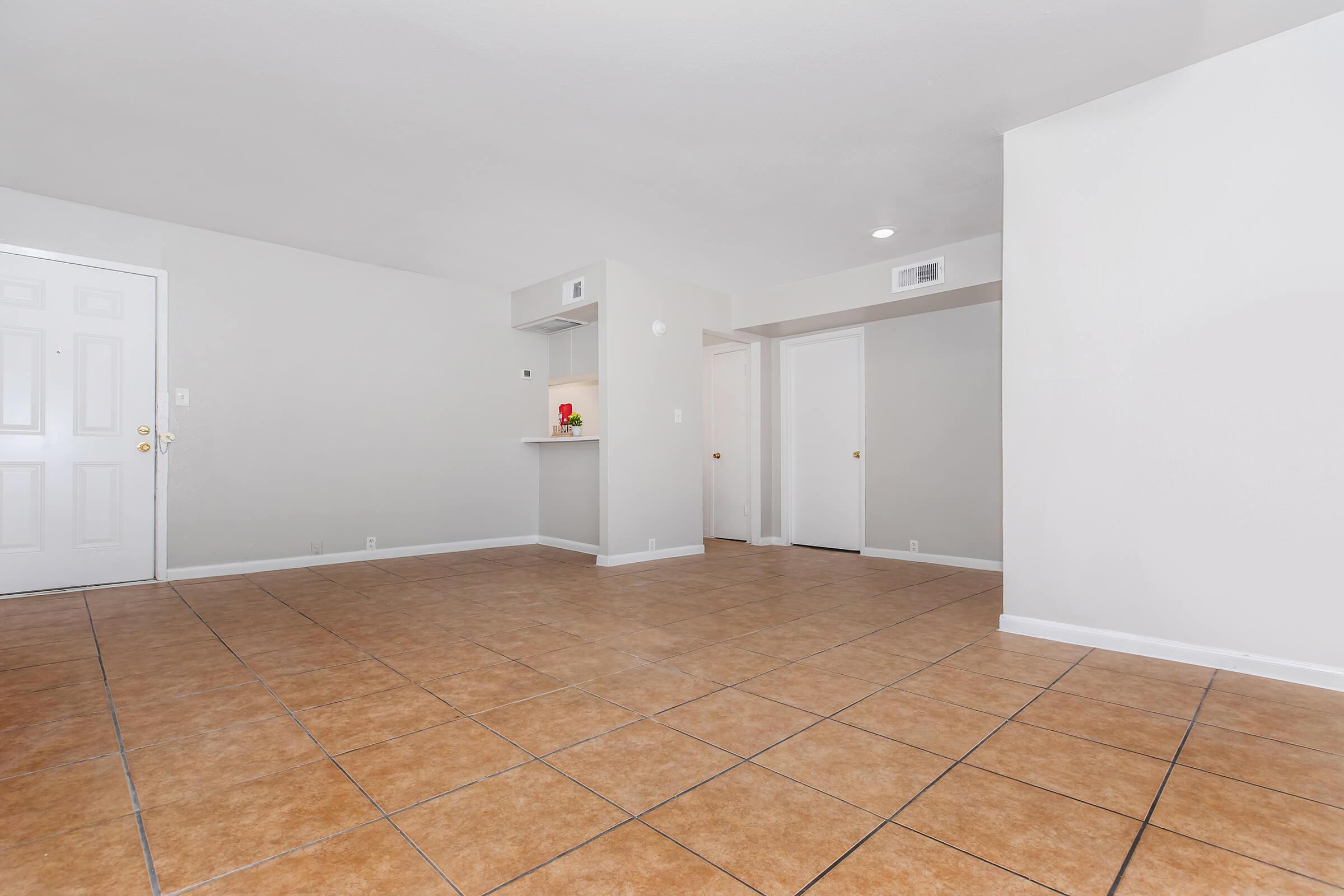 Interior of an empty apartment featuring beige walls and large brown tiled flooring. A doorway leads to a small kitchen area with a counter. Additional doors are visible, suggesting access to other rooms or storage. The space is well-lit and has a clean, minimalist aesthetic.