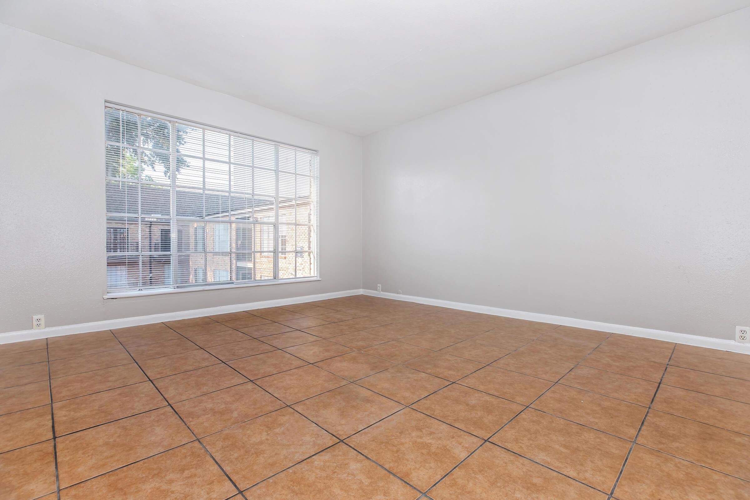 A spacious room with light gray walls and a large window featuring white blinds. The floor is covered in brown tiled flooring, creating an open and airy atmosphere. The room is empty, providing a blank canvas for decoration and furniture placement. Natural light comes in through the window.
