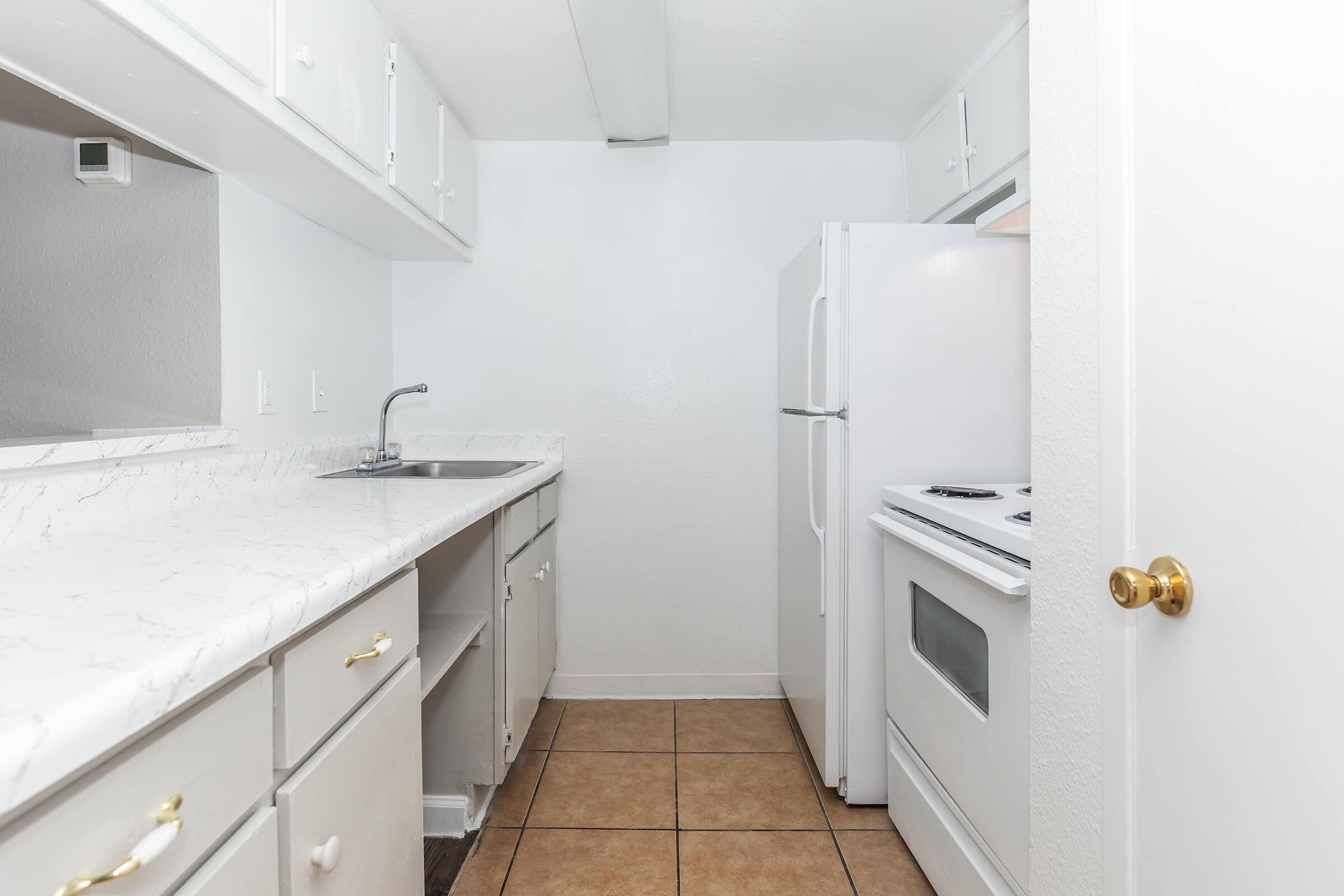 A small, modern kitchen featuring white cabinetry, a marble countertop, and light brown tile flooring. It includes a single sink, a white refrigerator, and a stove with an oven. The walls are painted white, enhancing the bright, clean appearance of the space.