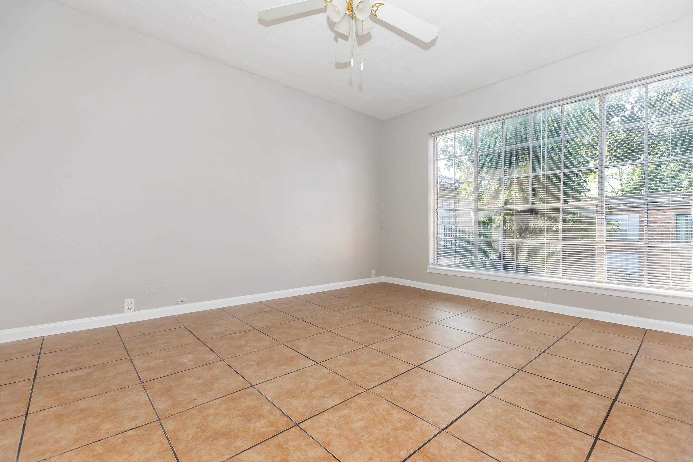 Empty room featuring beige tiled floor, light gray walls, and a large window with blinds. The ceiling has a ceiling fan with white blades, providing a bright and airy atmosphere. Natural light streams in through the window, enhancing the spacious feel of the area.