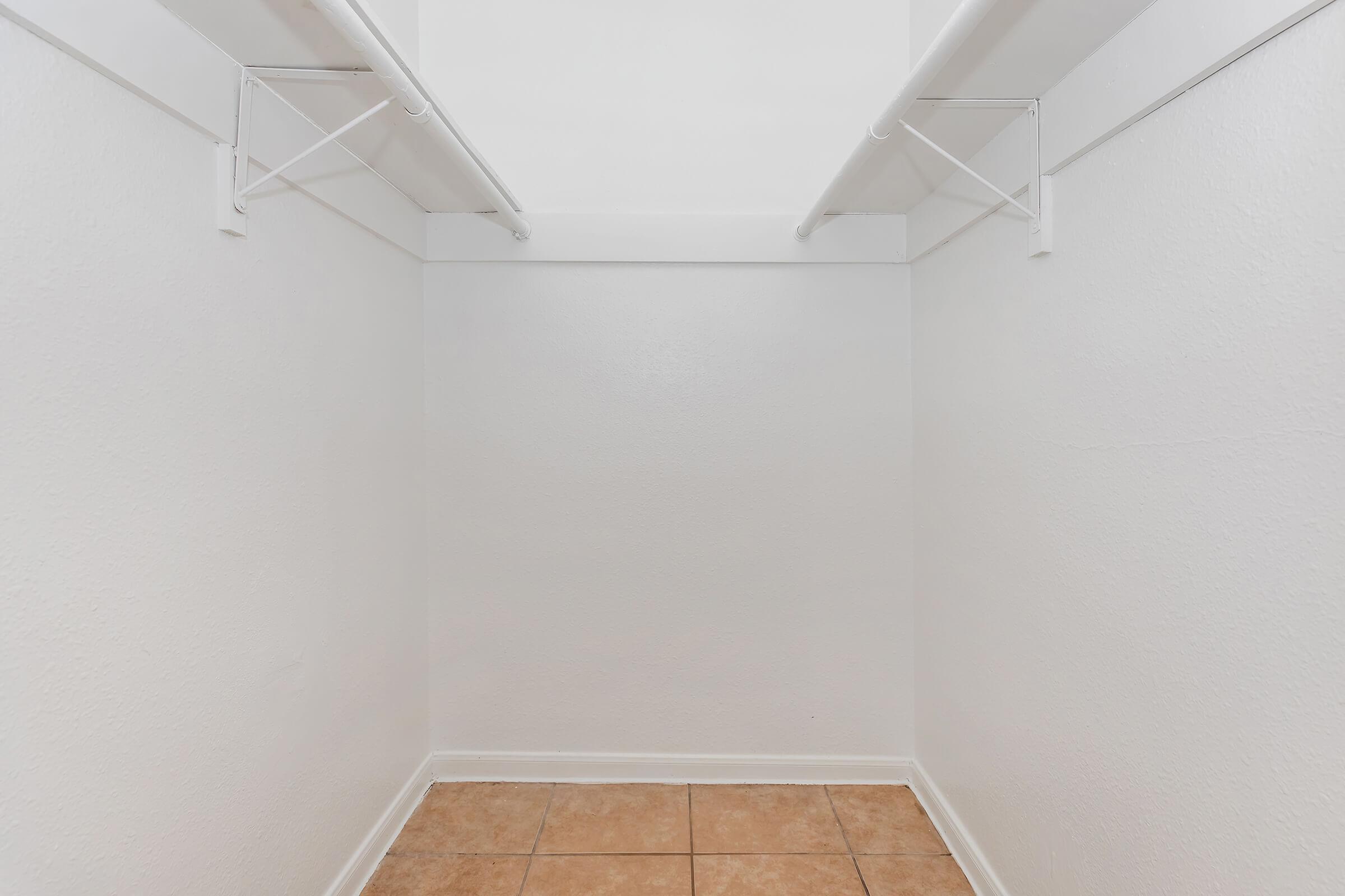 Empty closet with white walls and two metal hanging rods for clothes. The floor is tiled, featuring beige and light brown tiles. The overall space is well-lit and minimalistic, creating an open and airy feel.