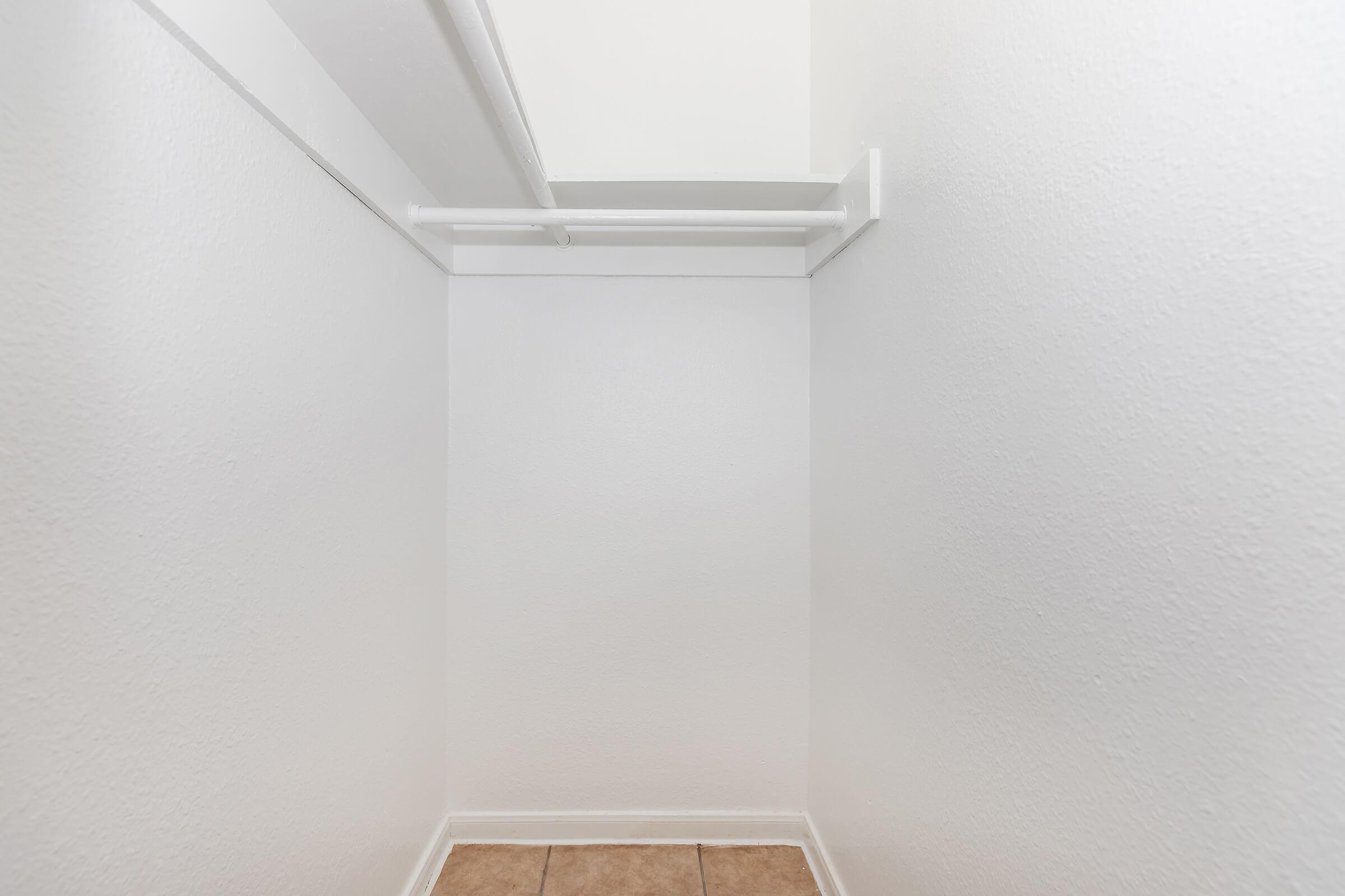 A small, empty closet with white walls and a beige tile floor. There is a single white rod on one wall for hanging clothes, and the ceiling is not visible, creating a bright, open space.