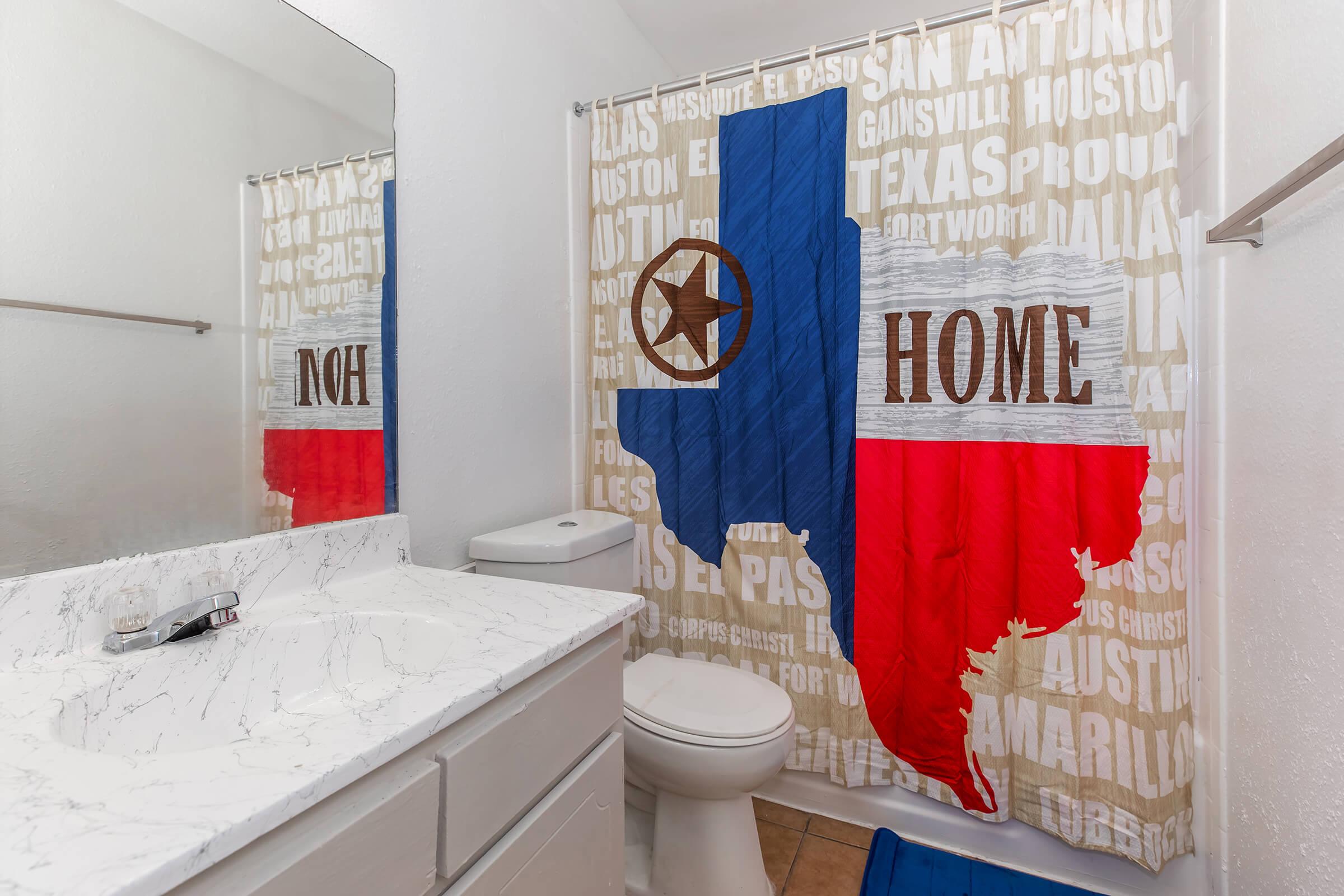 A bathroom featuring a shower curtain designed with the Texas flag, displaying the state outline and the words "HOME" and "NOH." The bathroom includes a white vanity with a marble countertop, a toilet, and a blue bath mat on the floor. The overall decor has a Texas theme.