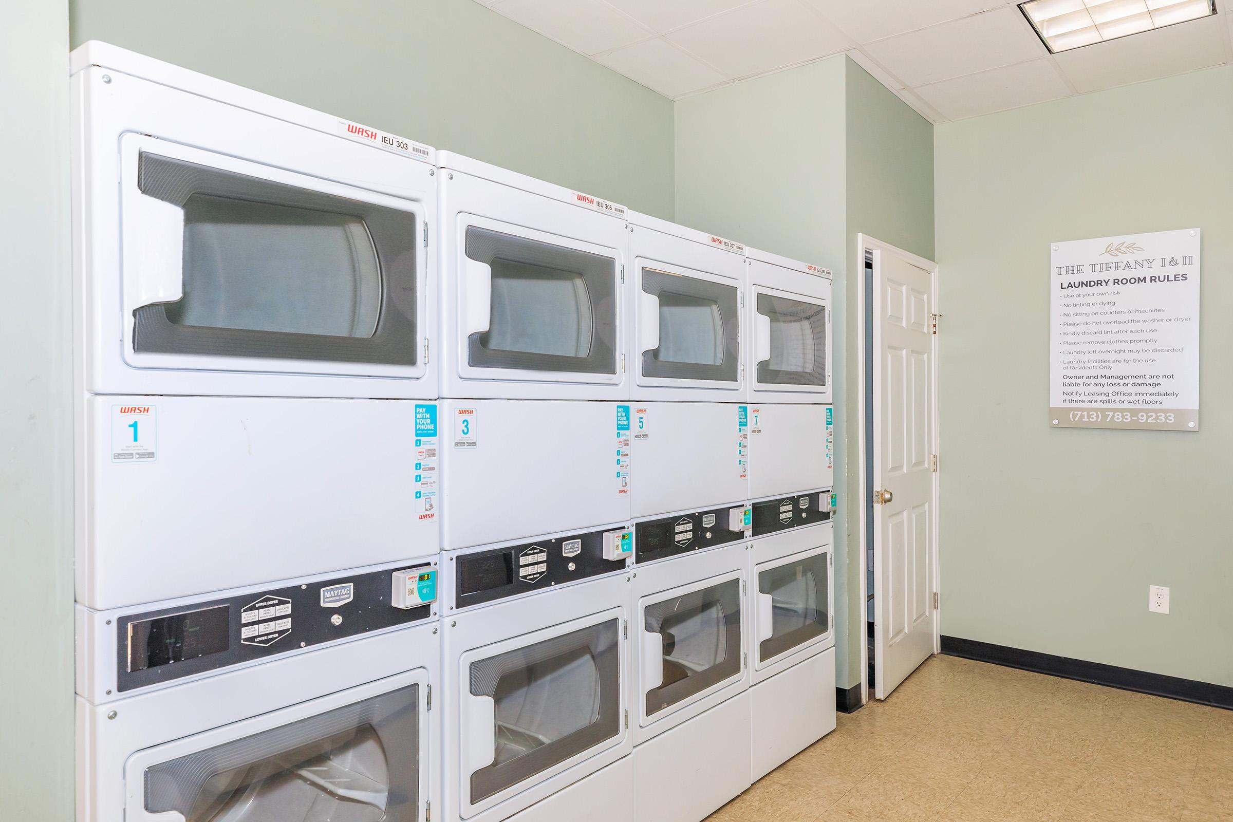 Rows of stacked dryers in a well-lit laundry room with a light green wall. A notice board is visible to the right, displaying information about laundry room rules and contact details. The flooring is a neutral color, contributing to a clean and organized appearance.