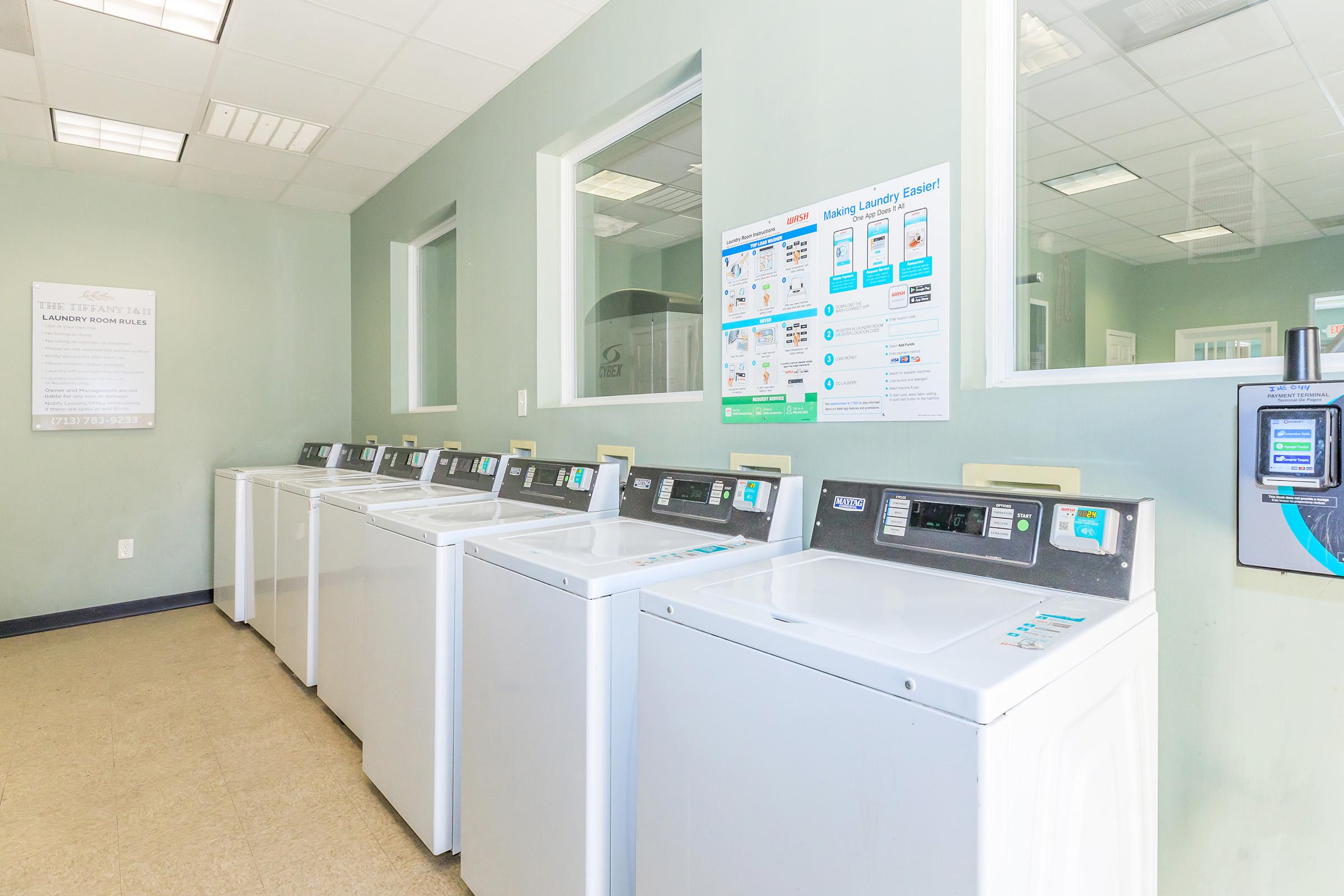 A bright and spacious laundry room featuring several white washing machines in a row. The walls are painted light green, and there are informational posters on the wall explaining laundry instructions. Large windows allow natural light to fill the space.