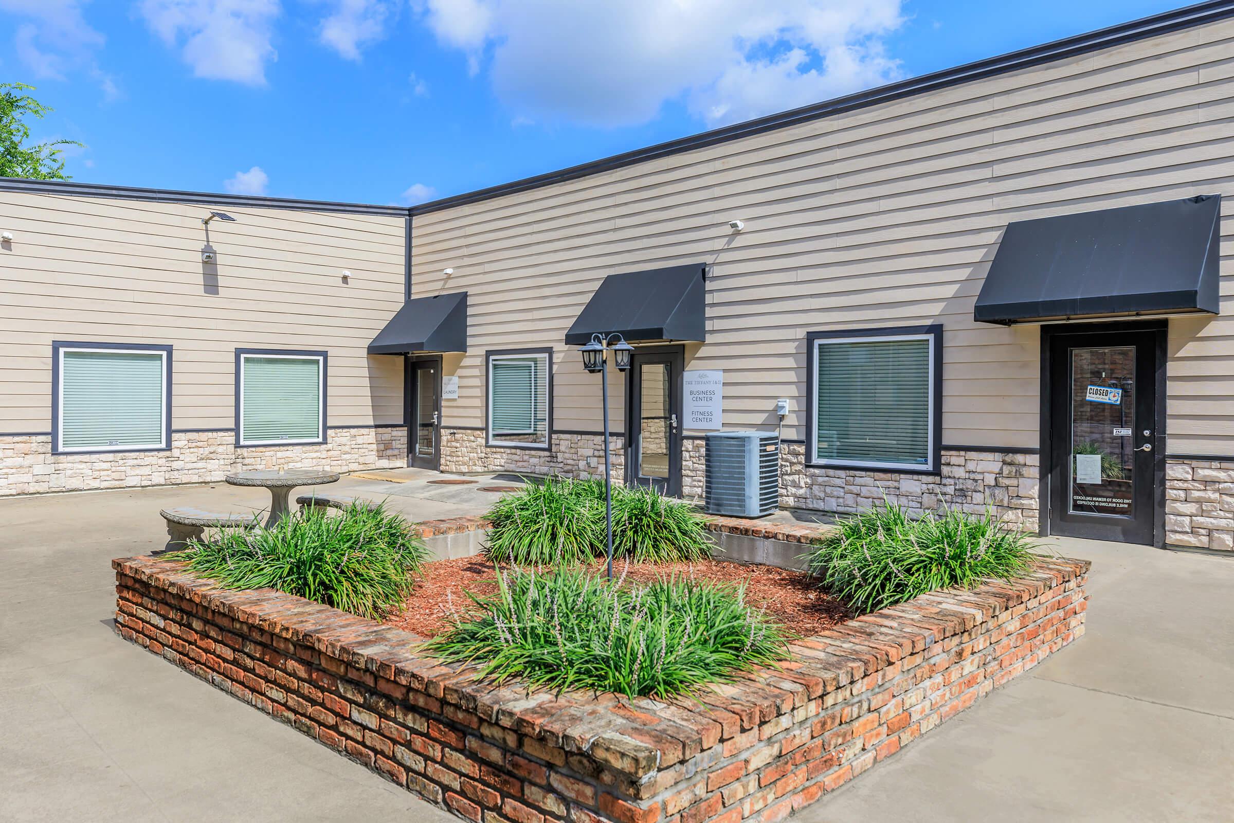 A modern commercial building featuring a landscaped courtyard with greenery and a brick planter. The facade has a combination of light-colored siding and stonework, with black awnings over the windows and doors. There are two entrance doors visible, and a bench is placed in the courtyard.