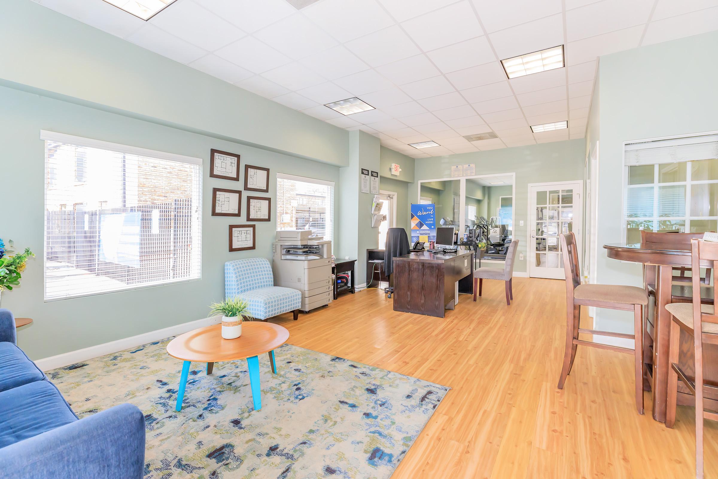 A bright and welcoming office space featuring a blue couch, a round coffee table with a small plant, and a work desk. There are framed pictures on the wall, a dining table with chairs, and large windows allowing natural light to fill the room. The flooring is wood laminate, creating a cozy atmosphere.