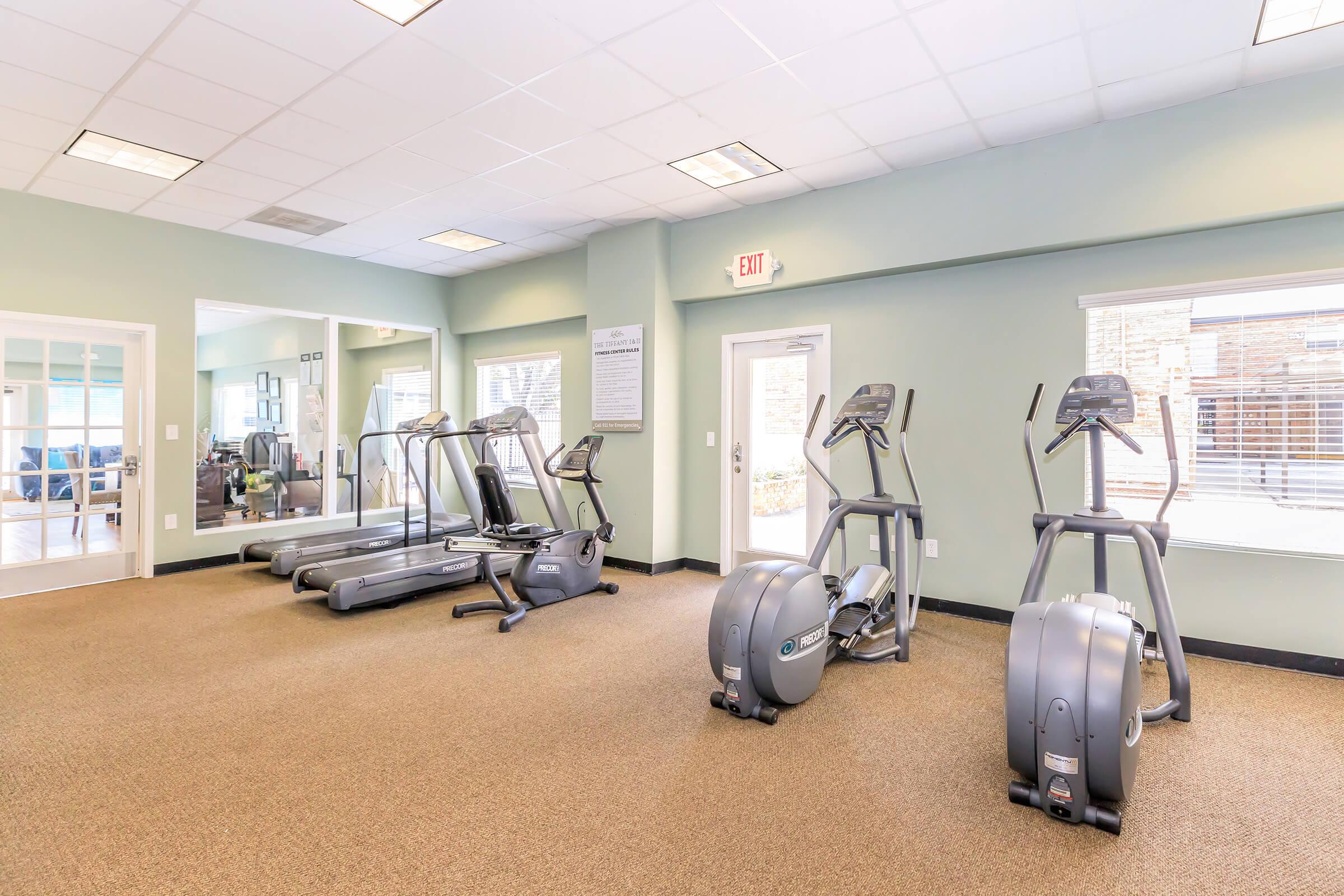 A well-lit gym area featuring exercise equipment, including multiple treadmills, ellipticals, and weight benches. The room has large windows allowing natural light, a soft carpeted floor, and mirrors on one wall, creating an inviting space for workouts.