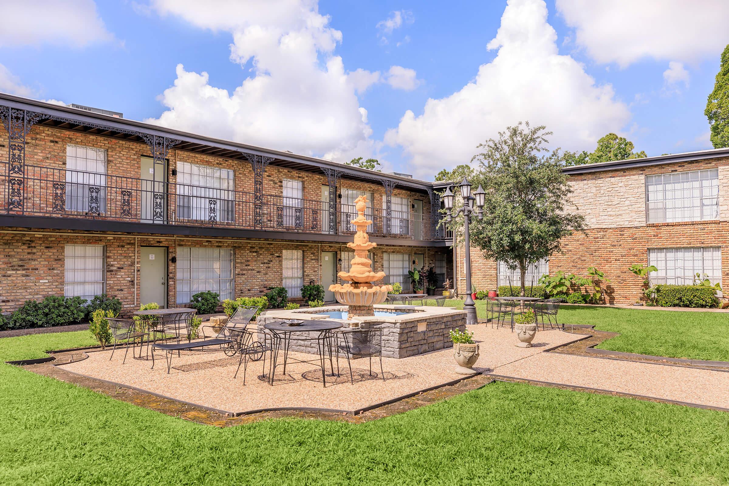 A charming courtyard featuring a central fountain surrounded by decorative potted plants and patio furniture. The area is bordered by two-story brick buildings under a bright, partly cloudy sky, creating a welcoming outdoor space for relaxation.