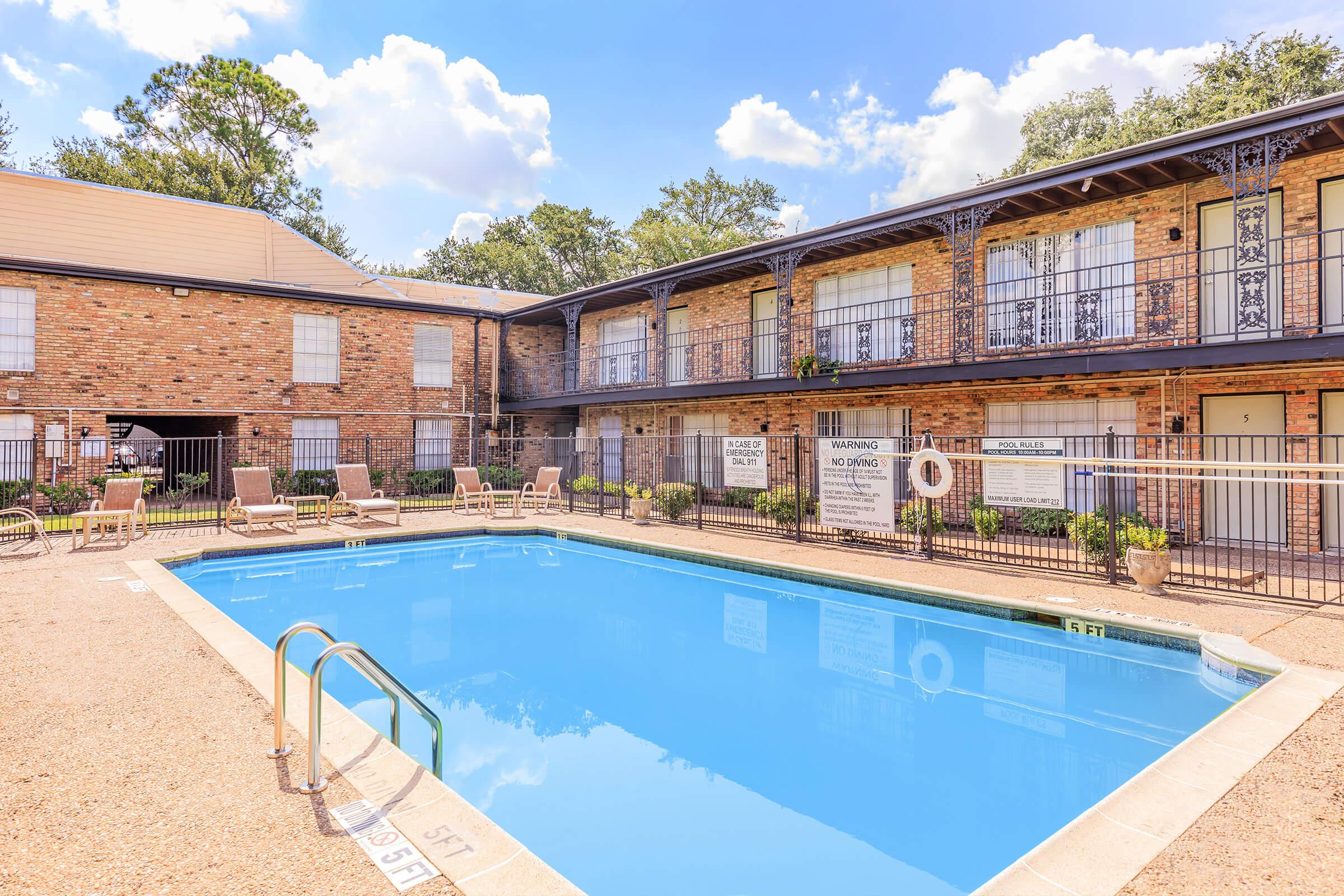 A clear blue swimming pool surrounded by a deck with lounge chairs, situated in a courtyard with brick apartment buildings. The buildings feature balconies and several signs around the pool area. Lush trees are visible in the background under a bright, partly cloudy sky.