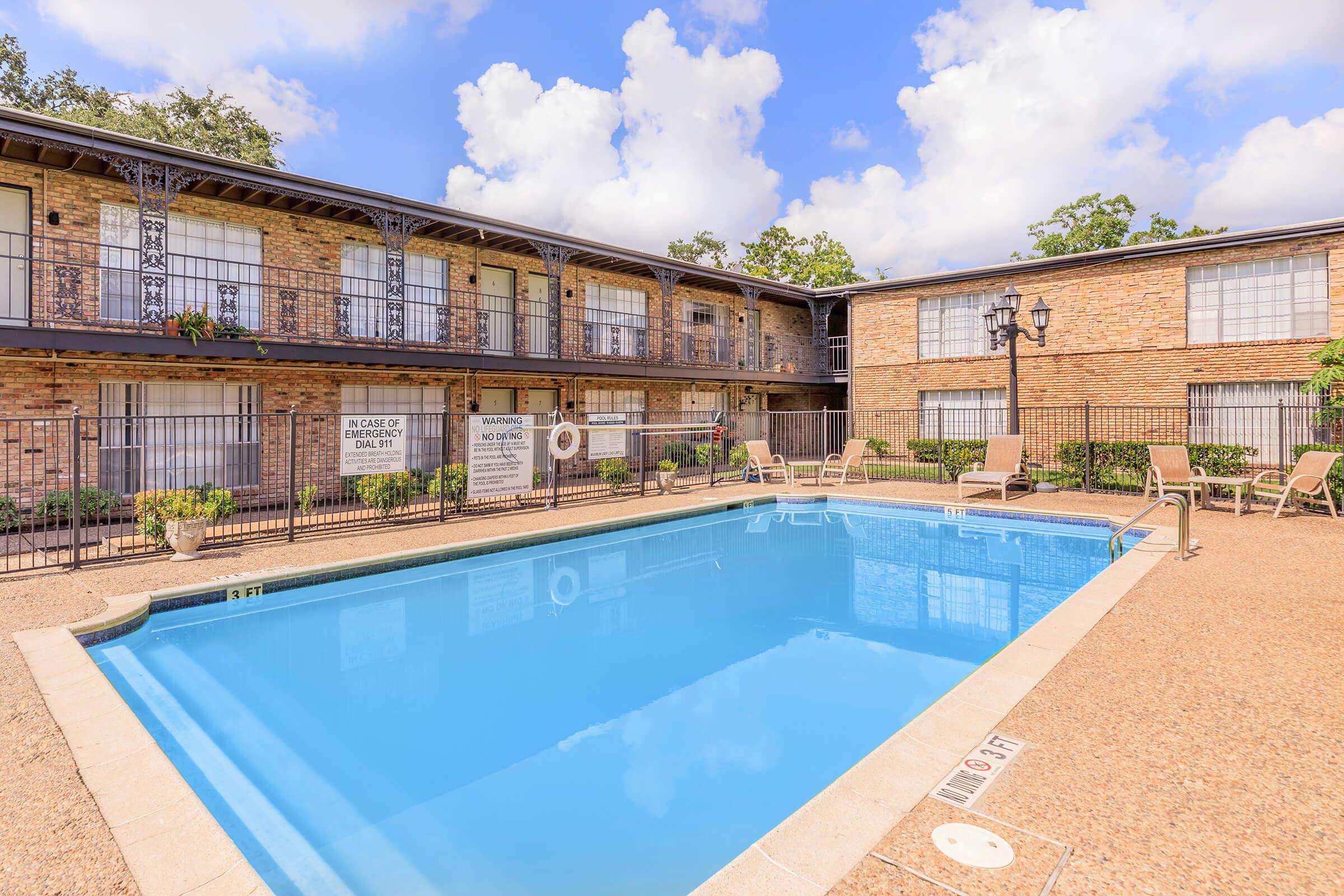 A clear blue swimming pool surrounded by a courtyard with brick buildings. Lounge chairs are placed around the pool area, and a lamppost adds to the ambiance. Lush greenery is visible, with clouds in a bright blue sky above. Signs are posted near the pool, indicating rules or information.