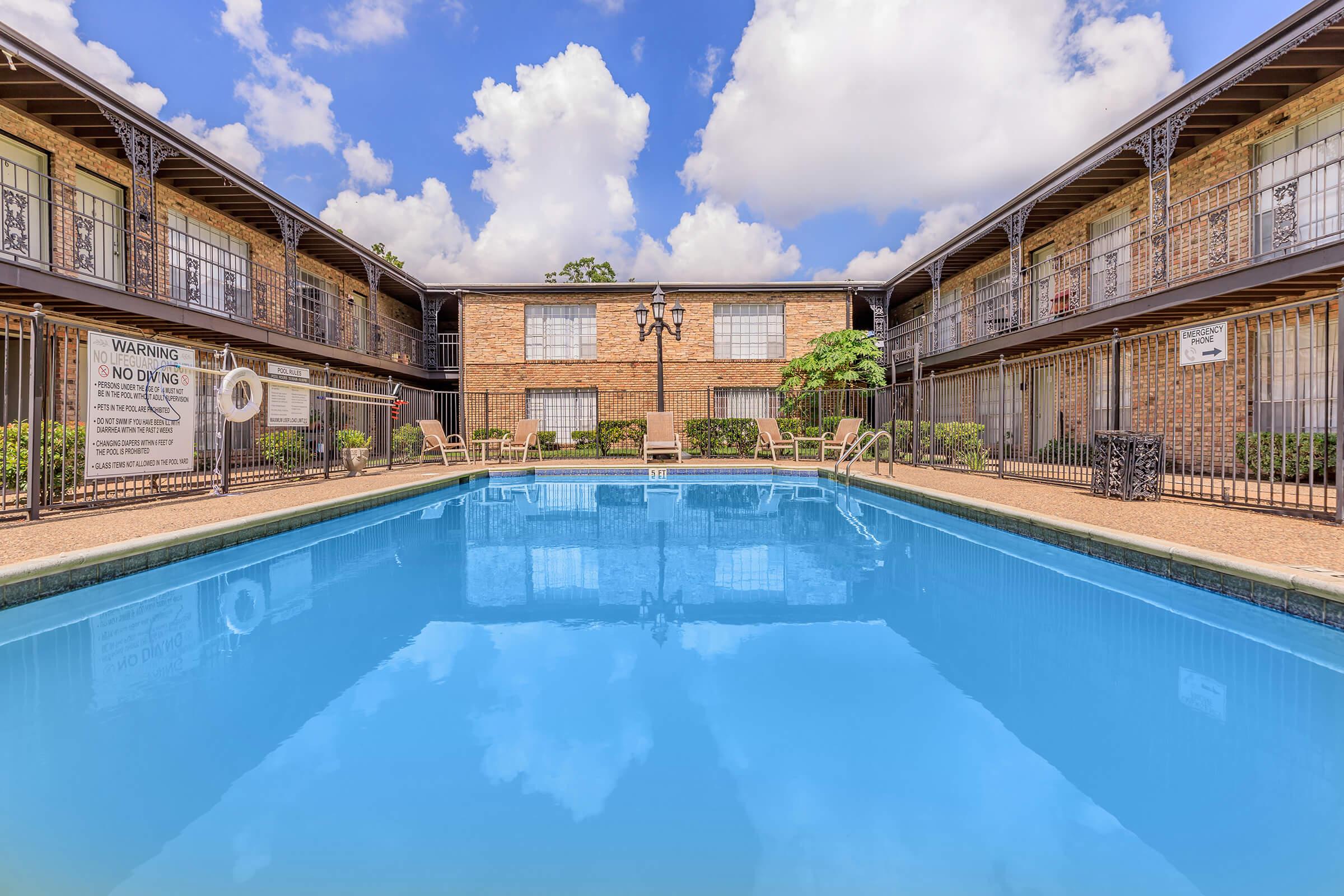 A clear blue swimming pool surrounded by a brick apartment building. The pool area features lounge chairs and is framed by trees and clouds in the sky. Signs are posted nearby with a warning about no diving. The scene conveys a serene and inviting atmosphere.