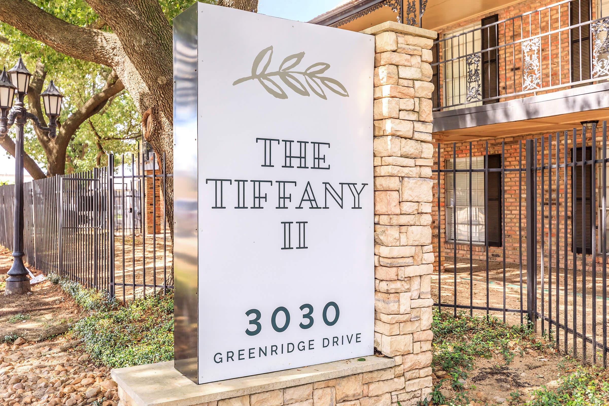 Sign for "The Tiffany II" located at 3030 Greenridge Drive. The sign features a minimalist design with a leaf emblem above the text. Surrounding the sign are trees and a brick building, with decorative lamp posts visible in the background.