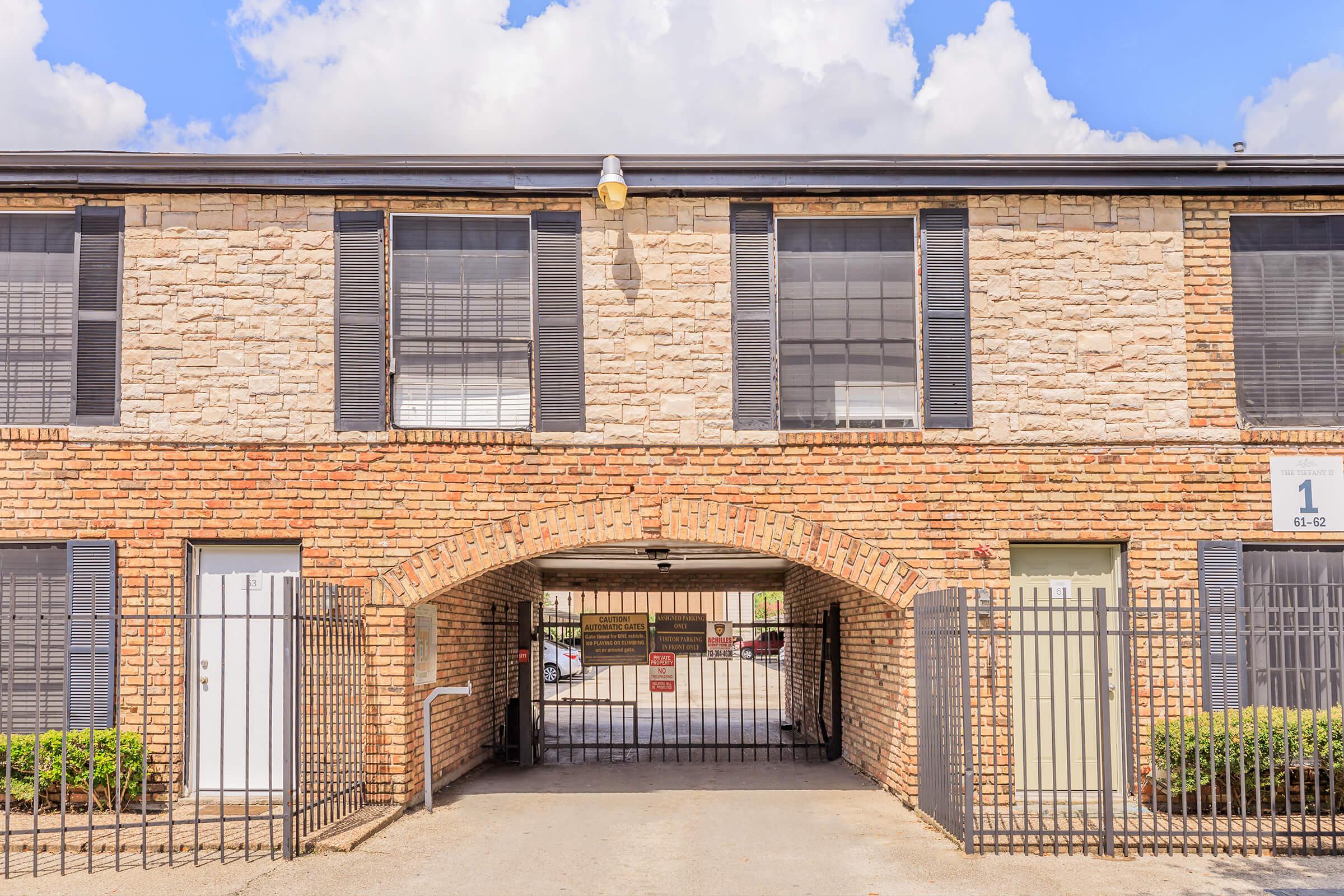A multi-story building with a sandstone facade and black shutters. The entrance features a gated archway leading to a parking area. Two doors are visible on either side, and the building is set against a backdrop of partly cloudy skies. Lush greenery lines the ground in front.