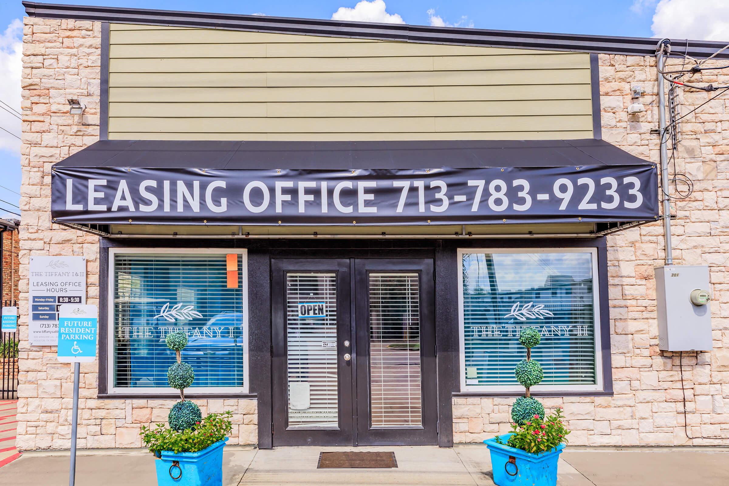 A leasing office building with a black awning displaying the phone number "713-783-9233." The entrance features double doors with an "OPEN" sign, large windows with blinds, and decorative potted shrubs on either side. The exterior is light-colored stone with a green upper section.