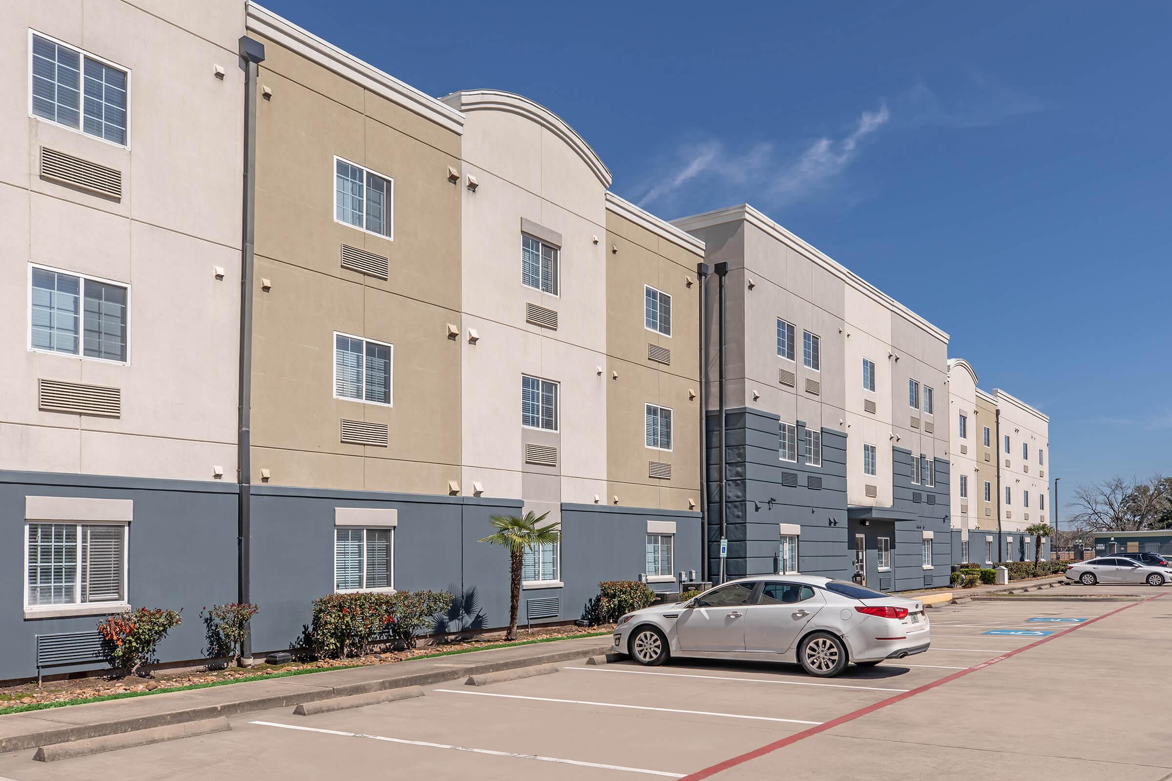 Exterior view of a multi-story hotel building with a combination of beige and gray siding. Several windows are visible along the facade, and a white sedan is parked in the foreground. The scene is set under a clear blue sky, with landscaped bushes lining the parking area.