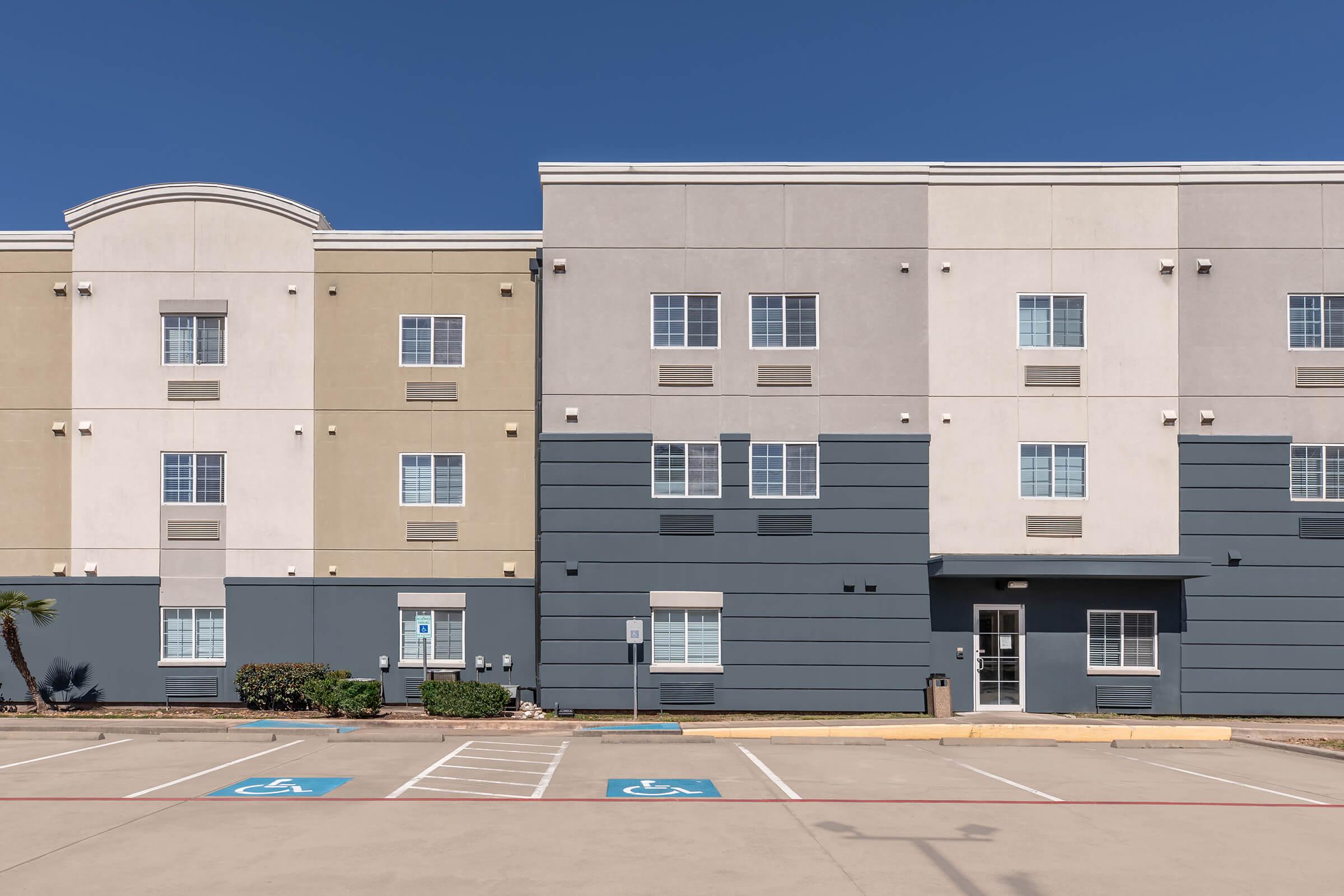 A modern, multi-story building featuring a combination of beige and gray exterior walls. It has multiple windows and a parking lot with accessible parking spaces marked. The scene is clear with a bright blue sky above.