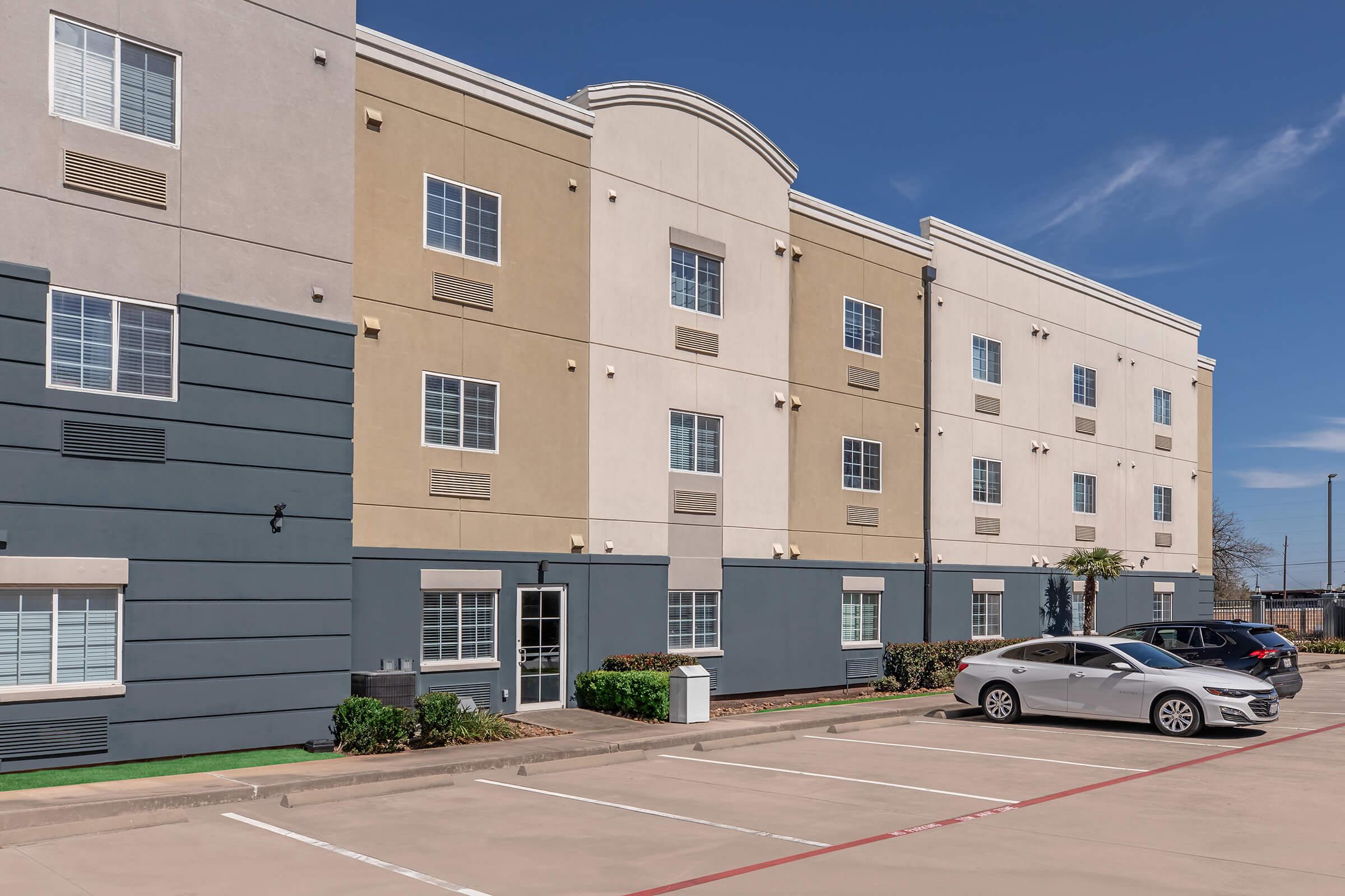 A modern, three-story hotel building with a mix of beige and gray exterior walls and multiple windows. In the foreground, there are parking spaces with two parked cars. The scene is set on a clear day with a blue sky.