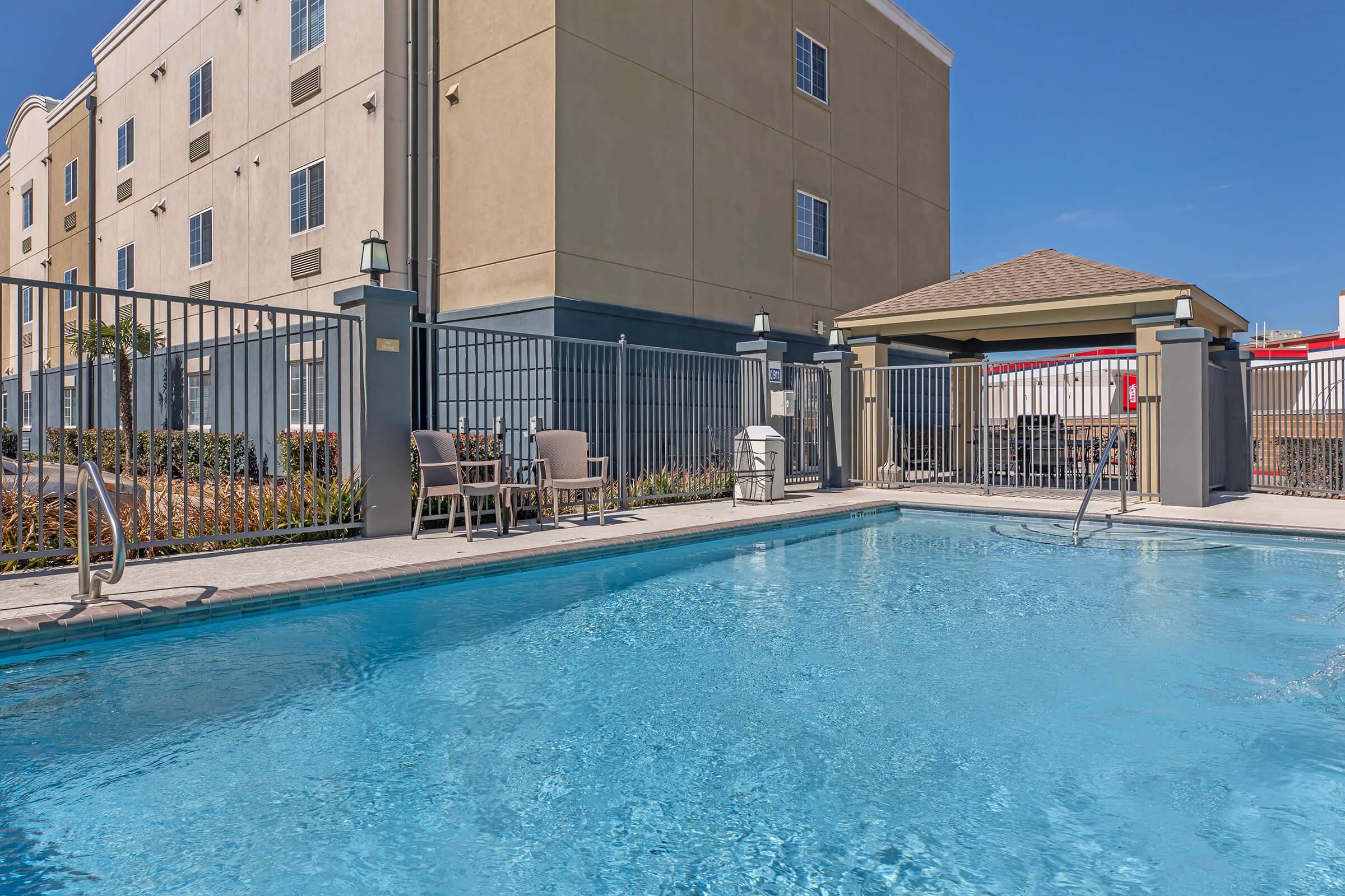 A clear swimming pool surrounded by a metal fence, with lounge chairs nearby. In the background, a beige building with several windows is visible under a bright blue sky. A poolside gazebo is also seen, creating a relaxing atmosphere.