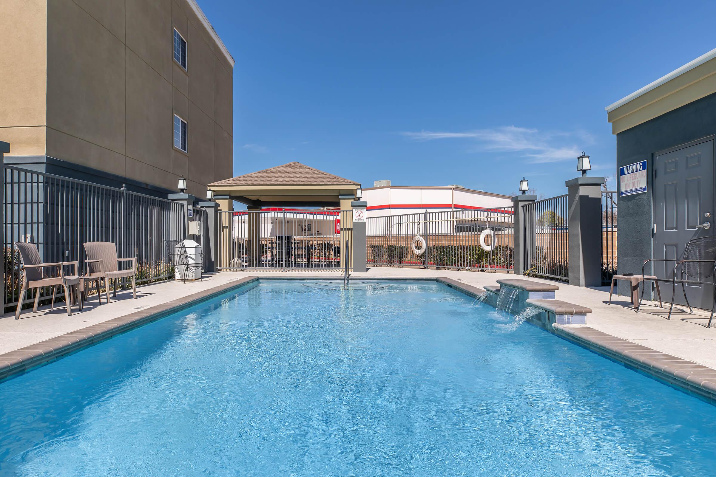 A clear blue swimming pool surrounded by a tiled deck, with lounge chairs on either side. A shaded area with a gazebo is visible in the background. The pool area is fenced, featuring safety floats and a sunny sky overhead.