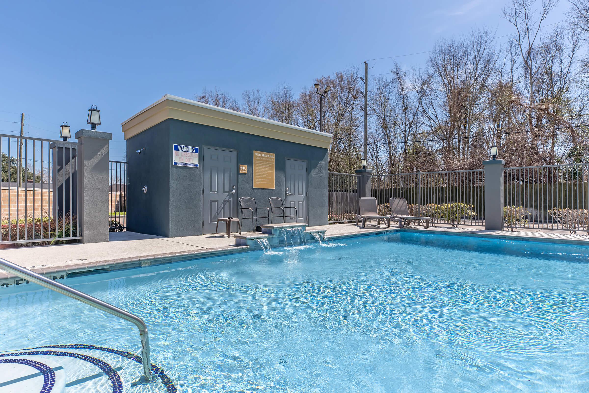 A swimming pool with a small building in the background. The pool features a spa-like area with water cascading from the building. Surrounding the pool are lounge chairs and a fence, with trees visible in the distance under a clear blue sky.