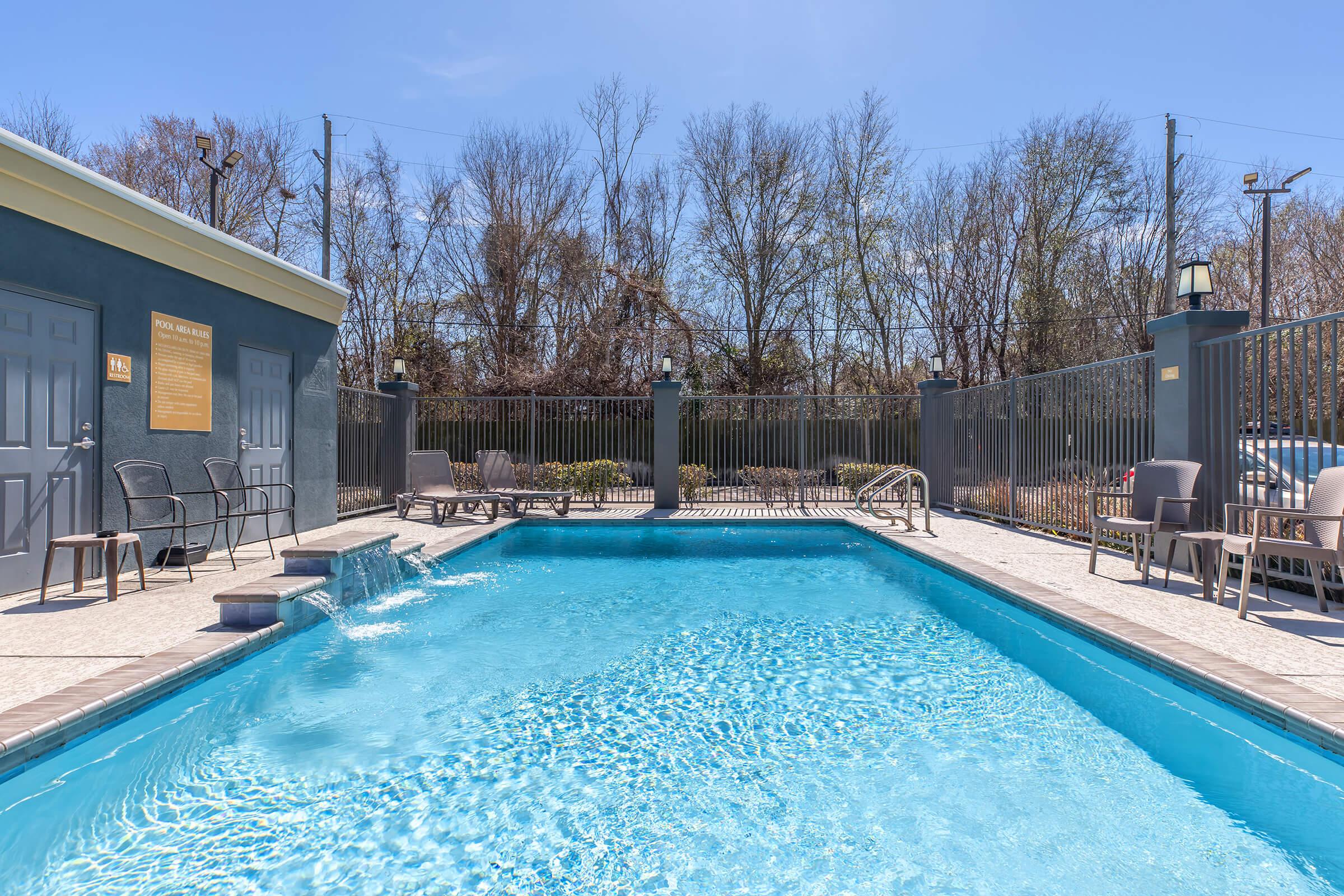 A clear blue swimming pool surrounded by a gated area. Lounge chairs are set up along the poolside. In the background, trees without leaves indicate an early spring or late winter season. The sky is bright and sunny, enhancing the inviting atmosphere.