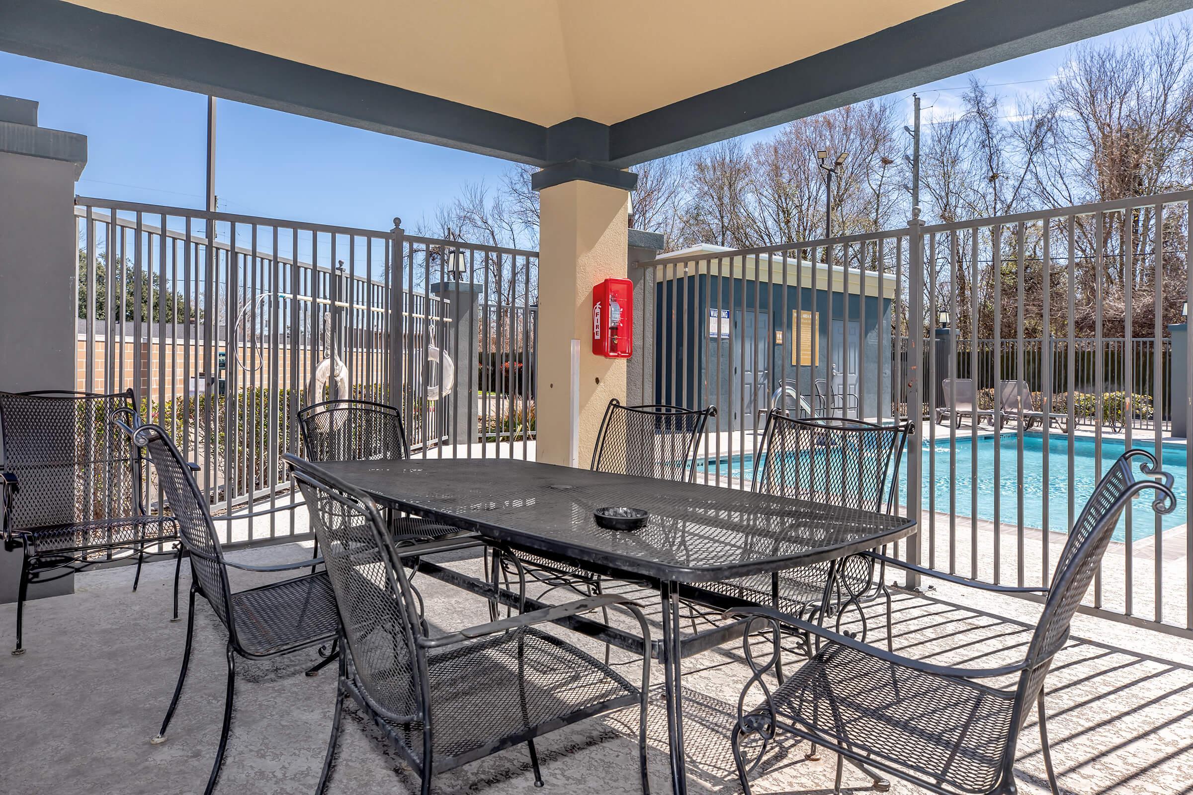 A shaded outdoor seating area featuring a large black metal table surrounded by chairs, with a view of a fenced swimming pool in the background. The scene conveys a relaxing atmosphere, ideal for gatherings. A red emergency phone is mounted on the wall nearby.