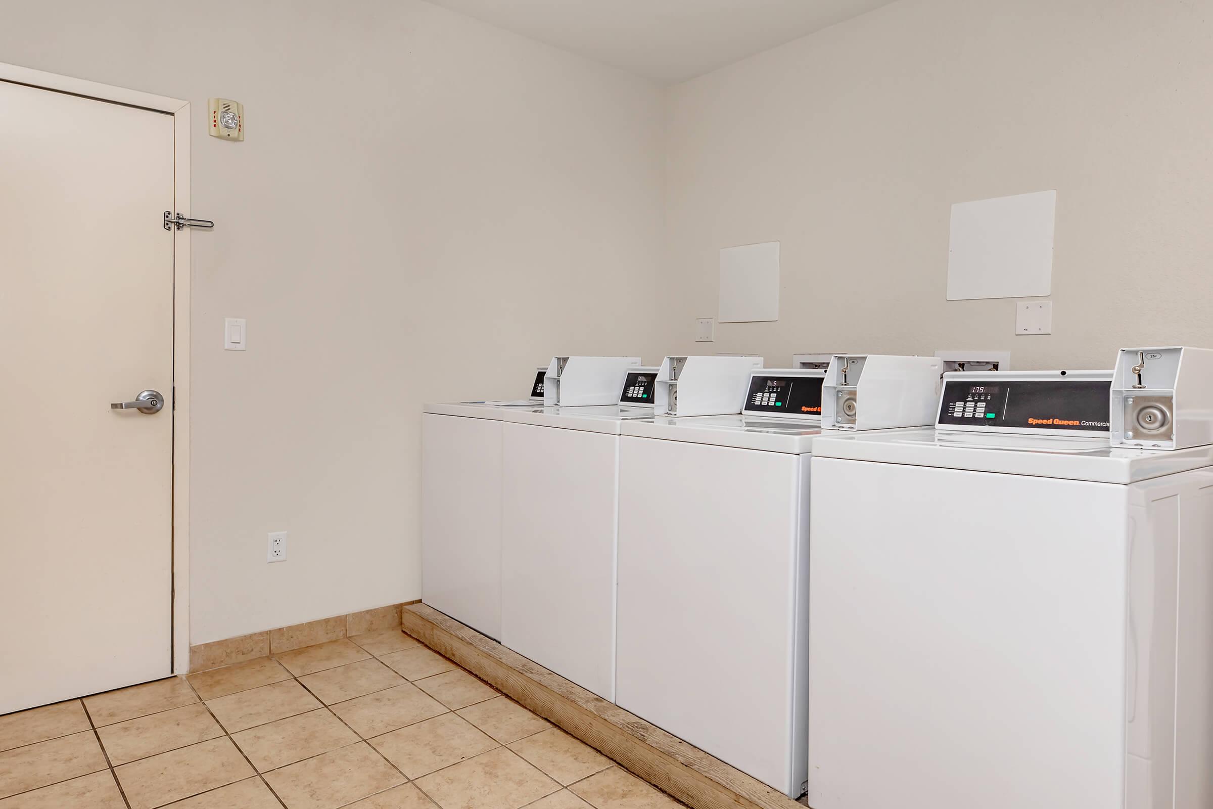 A laundry room featuring three white washing machines lined up against a wall, with a beige tiled floor. There is a door on the left side of the image. The walls are plain and light-colored, creating a clean and simple environment.