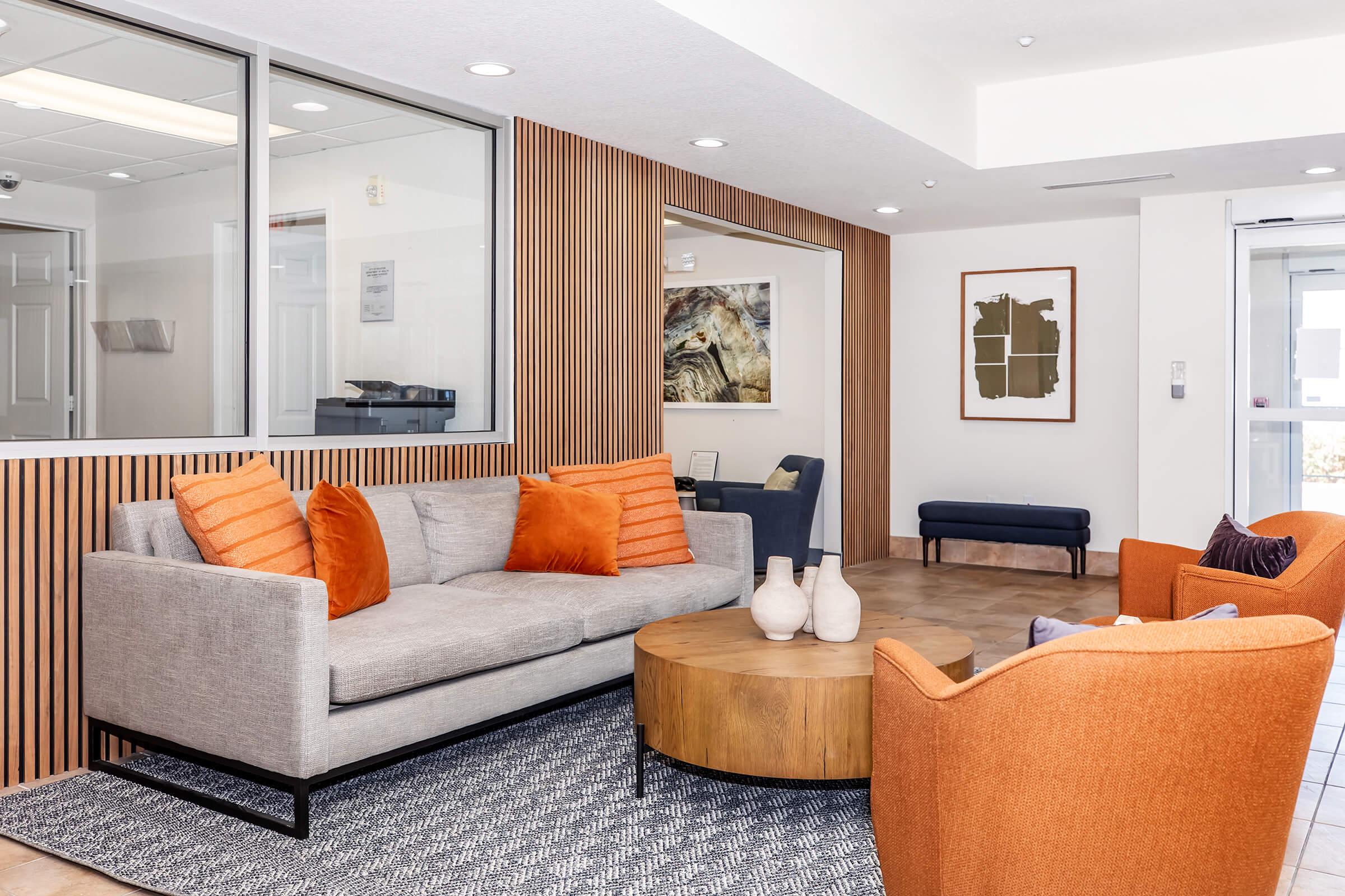 A modern lounge area featuring a light gray sofa with orange pillows, an orange accent chair, and a round wooden coffee table. The space has a textured wall in warm wood tones, a patterned rug, and contemporary artwork on the wall. Large windows allow natural light into the room.