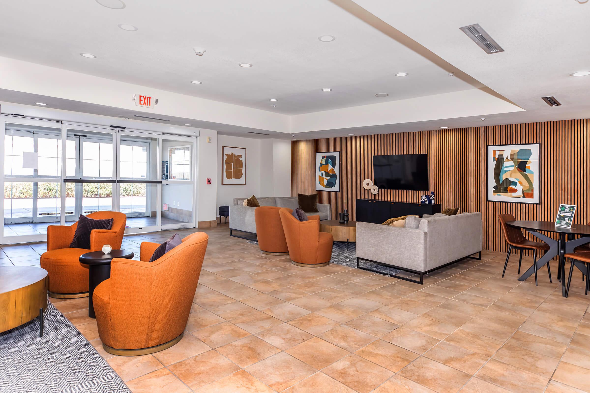 A modern lounge area featuring comfortable orange chairs, gray sofas, and a large flat-screen TV mounted on a wooden wall. The space includes a coffee table and decorative artwork on the walls, with large windows allowing natural light. The floor is made of light-colored tiles, creating a welcoming atmosphere.