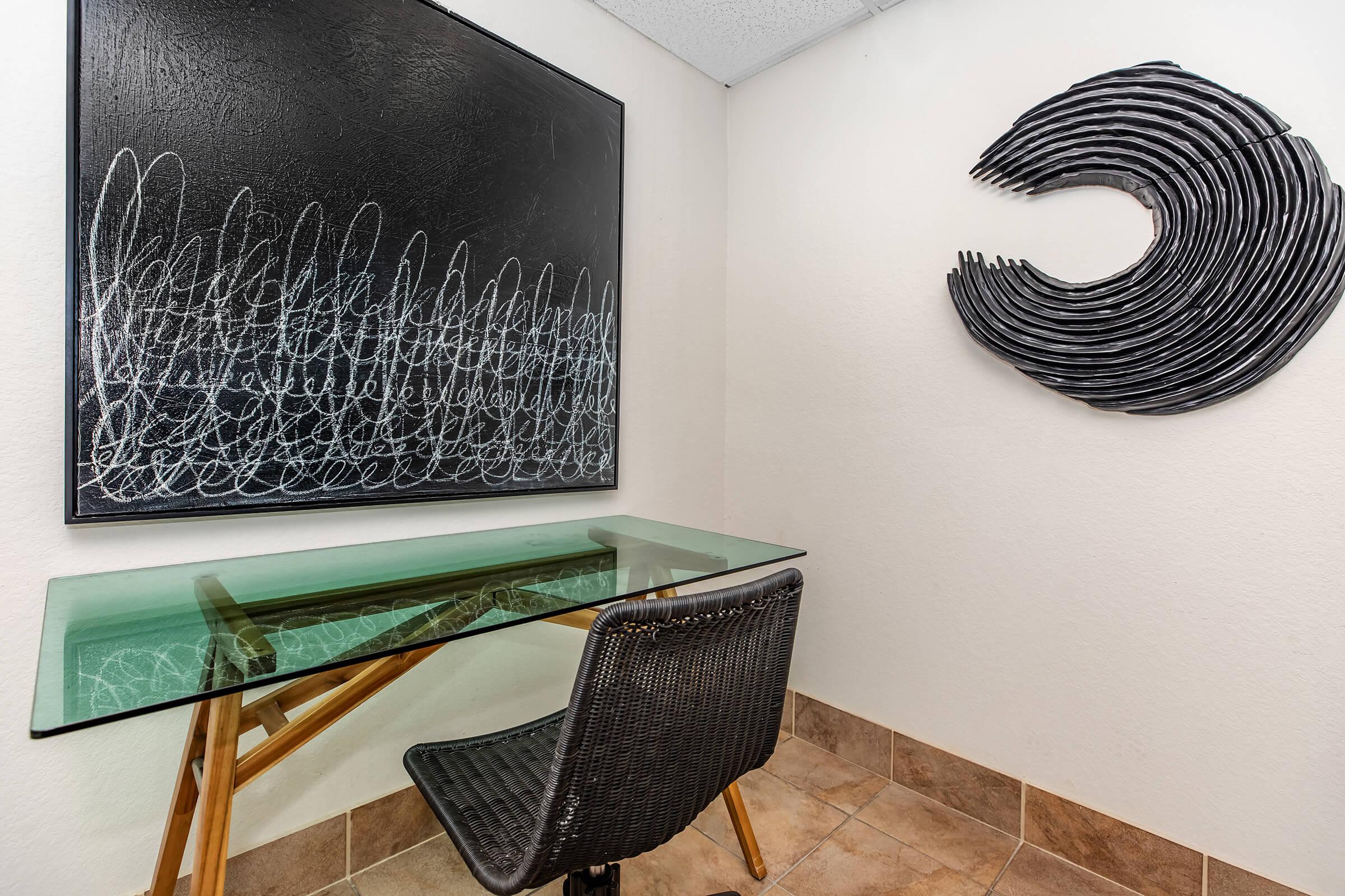 A small workspace featuring a glass-top table and a woven chair. The wall behind displays a large abstract black artwork with fluid white lines, and to the side, there is a curved black sculpture that resembles a wave. The floor is tiled, contributing to a minimalist aesthetic.