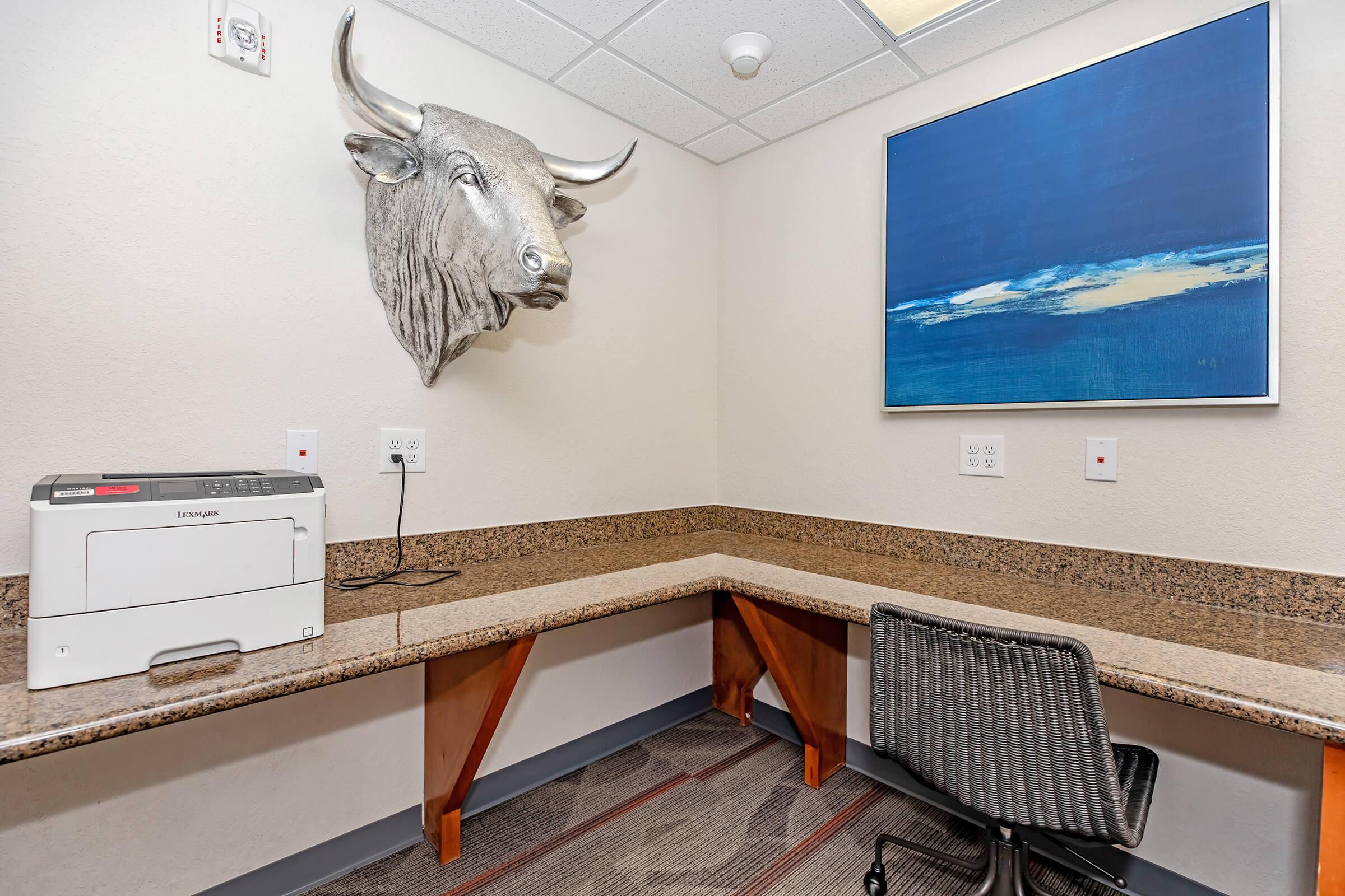 A small office space featuring a mounted bull's head on the wall. There's a printer on a wooden countertop, with a modern black office chair nearby. A blue abstract painting hangs above, and the walls are light-colored with electrical outlets visible. The overall atmosphere is professional and minimalist.