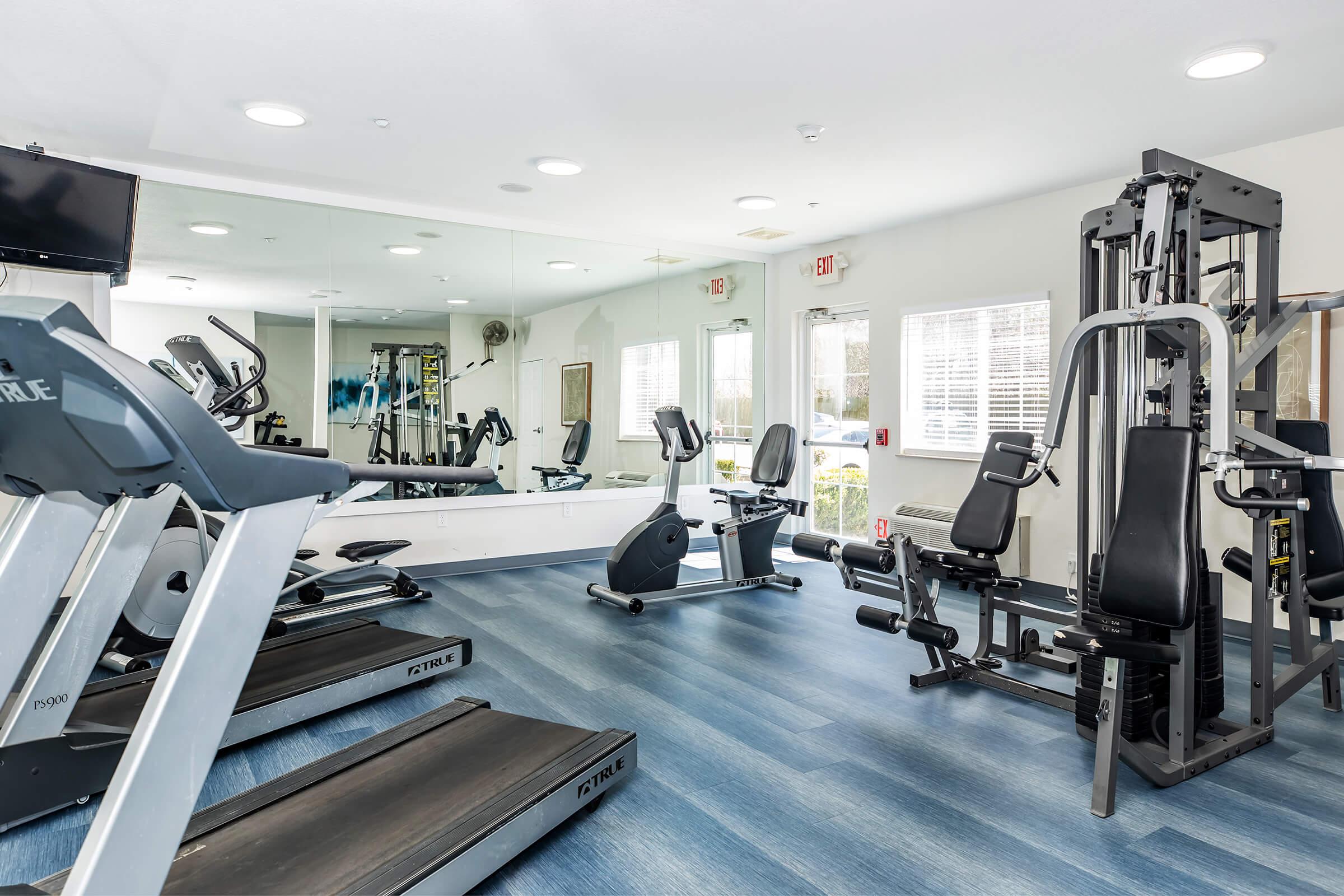 A well-lit gym featuring various exercise equipment, including treadmills and a multi-station weight machine. Large windows allow natural light to fill the space, and mirrors are mounted on one wall, enhancing the workout environment. Blue flooring complements the modern design.