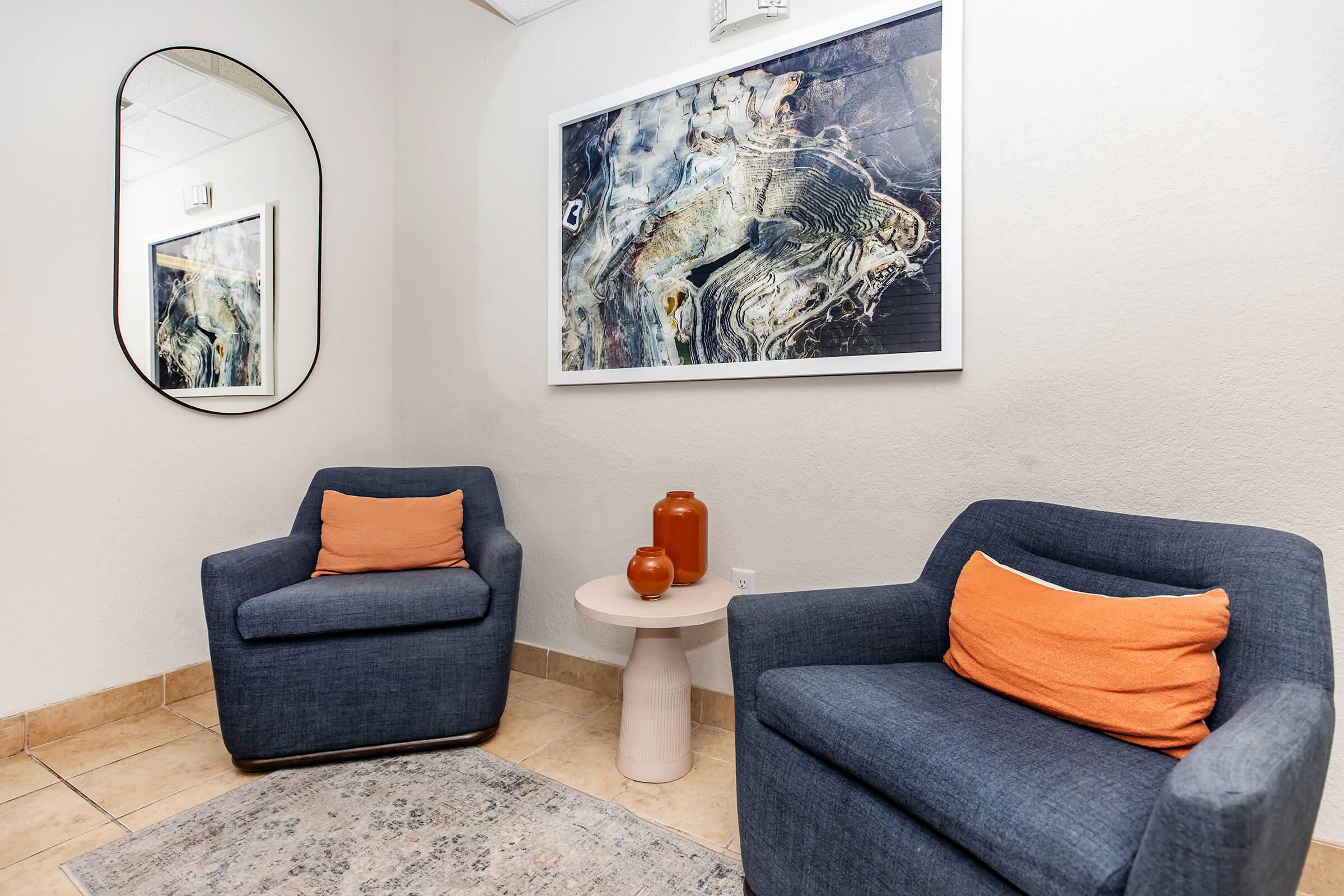 Two blue armchairs with orange cushions are placed in a cozy corner. A round, light-colored side table sits between them, topped with a small orange vase and a decorative bowl. The wall features a large framed artwork with abstract patterns, and a circular mirror reflects the space. The floor is tiled, and the overall atmosphere is inviting and modern.