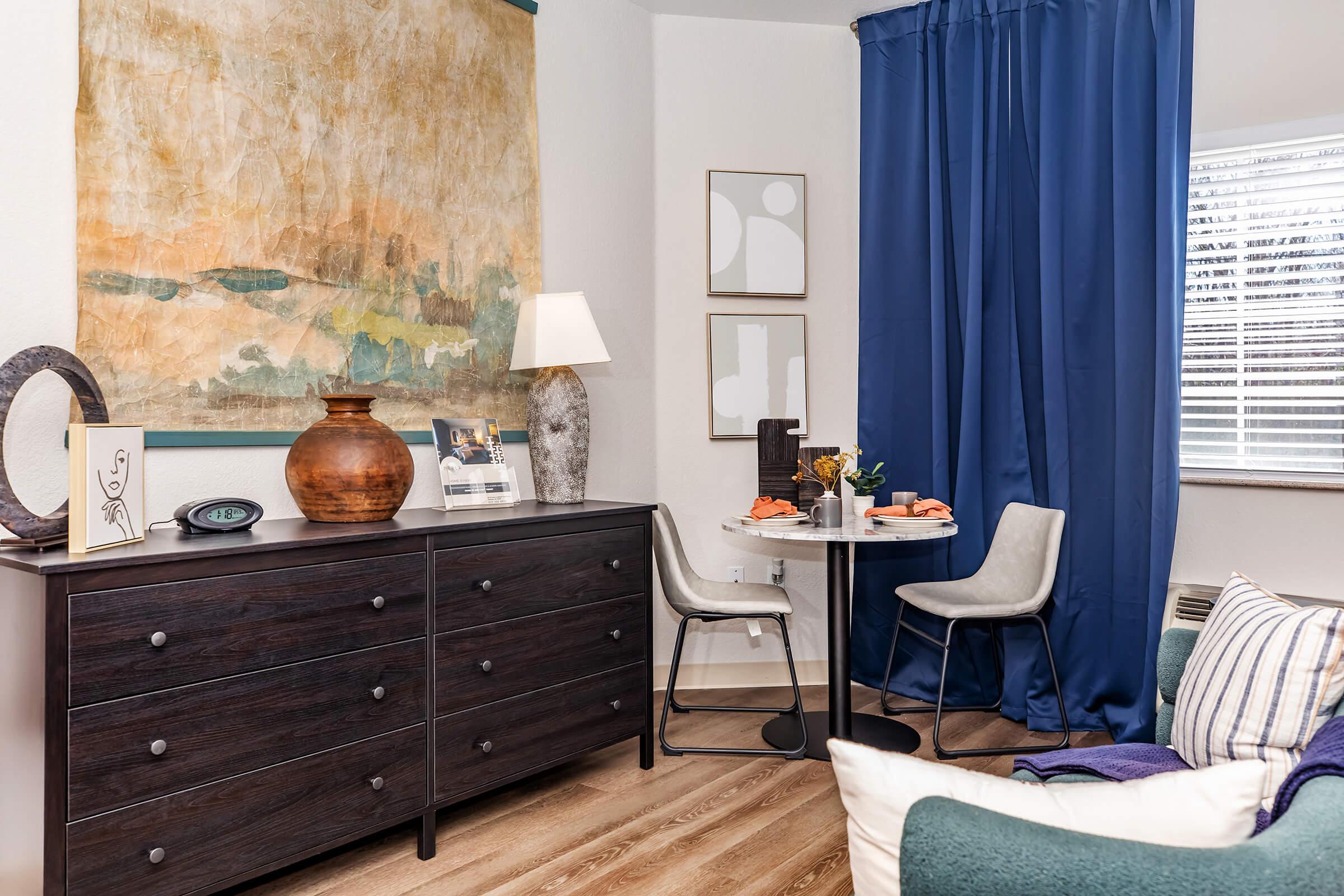 A cozy living space featuring a dresser with a decorative vase, a circular dining table with chairs, and a large abstract wall painting. There's a blue curtain by the window and a small centerpiece on the table. Warm wooden flooring complements the modern decor.