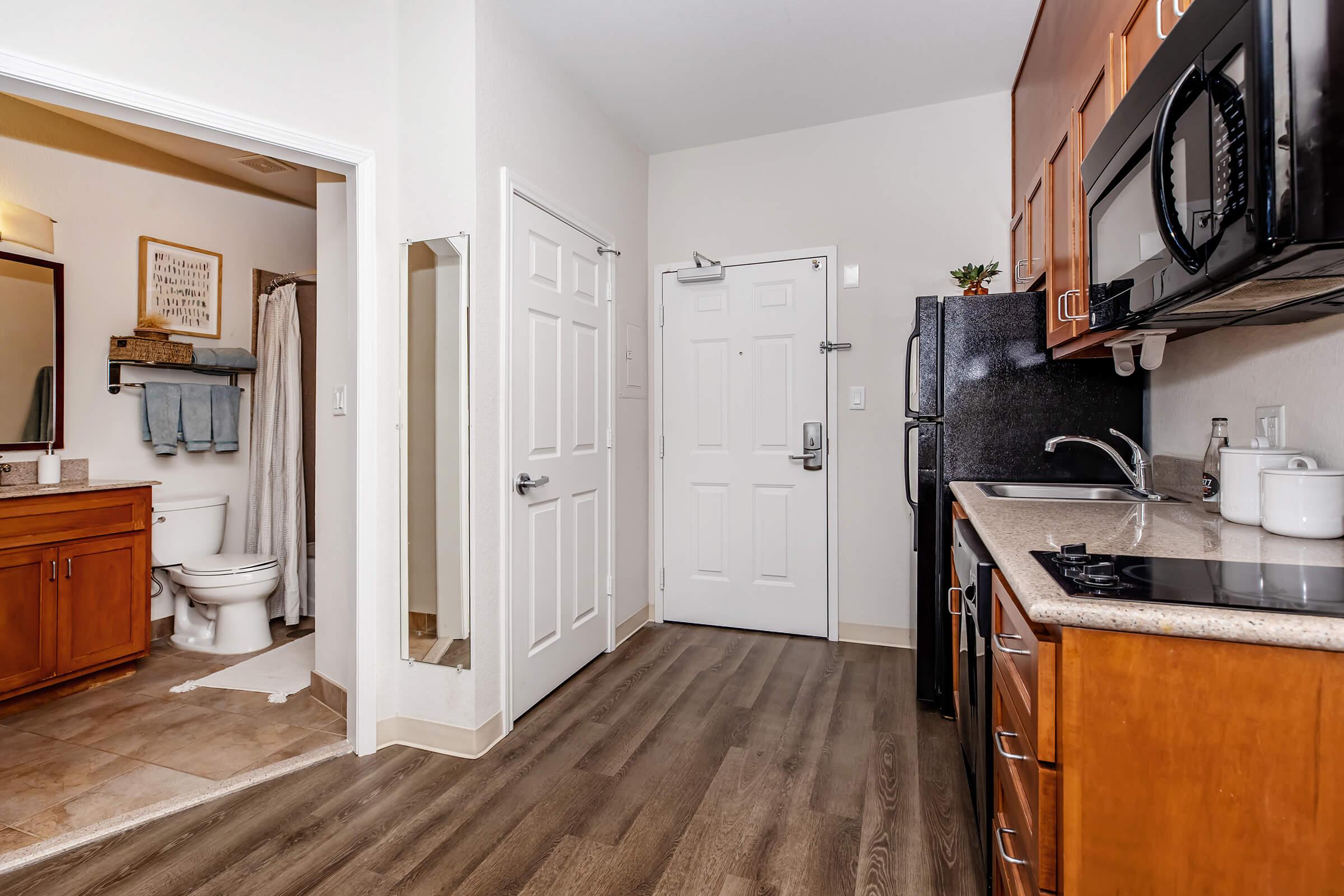 A modern kitchen and bathroom area with wooden cabinetry and appliances. The kitchen features a black refrigerator, microwave, and a stone countertop. A doorway leads to a bathroom with a toilet, and there is a mirror and towels visible. The walls are painted light colors, and there's a front door visible, adding to the welcoming feel.