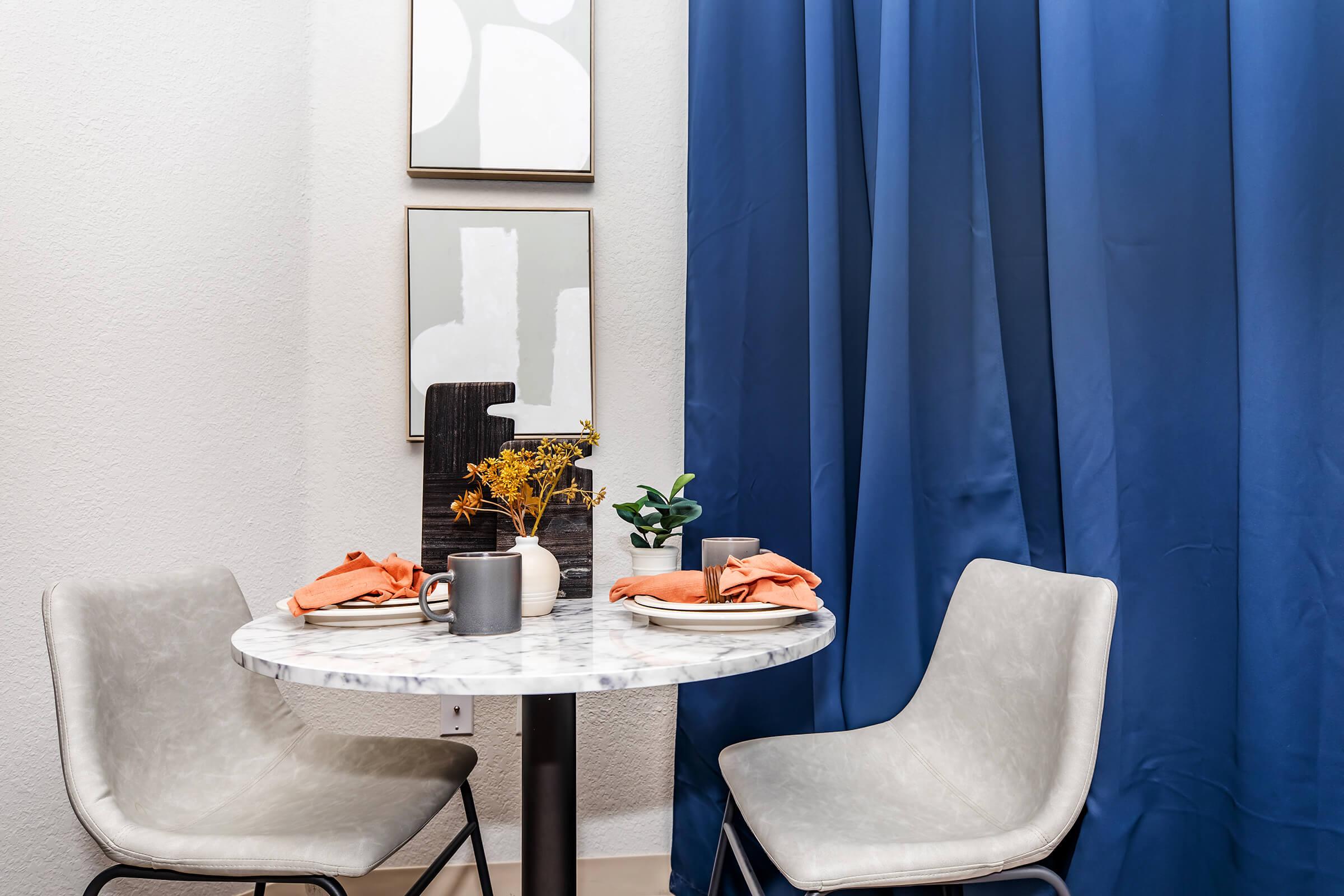 A cozy dining nook featuring a small round marble table with two gray chairs. The table is set with orange napkins, a minimalistic black centerpiece, and small decorative plants. Behind, blue curtains frame the scene, with two framed artworks hanging on the wall above the table.