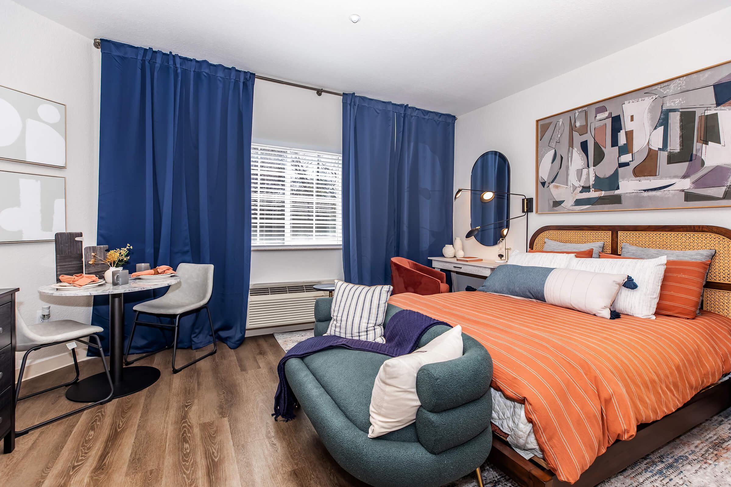 A stylish bedroom featuring an orange-striped bedspread, a blue accent chair, and a small round dining table with chairs. Large blue curtains frame the window, providing natural light. A piece of abstract art hangs on the wall, adding a modern touch to the cozy space. Warm wooden flooring enhances the inviting atmosphere.