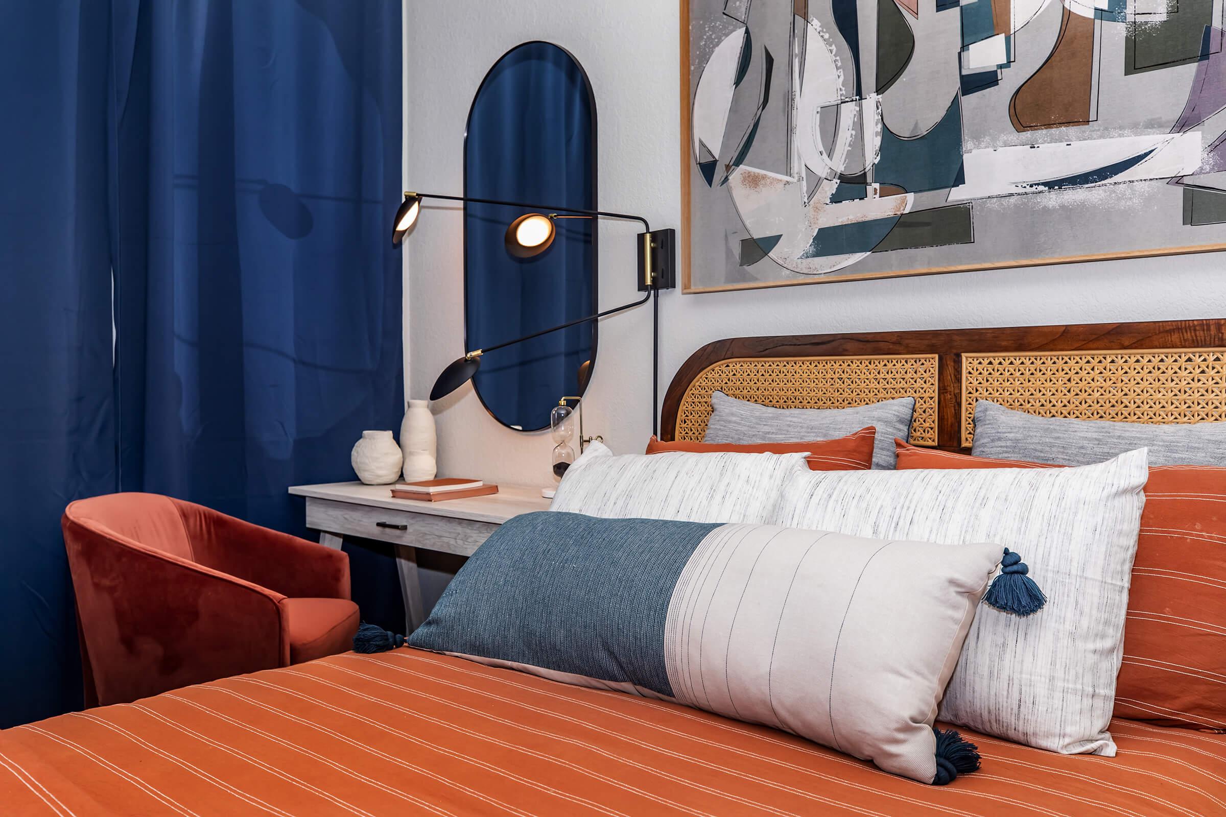 A cozy bedroom featuring a wooden bed frame with orange and blue striped bedding, decorative pillows, a plush rust-colored chair, a stylish desk with a mirror above, and navy blue curtains. Artwork with abstract designs hangs on the wall, creating a modern, inviting atmosphere.