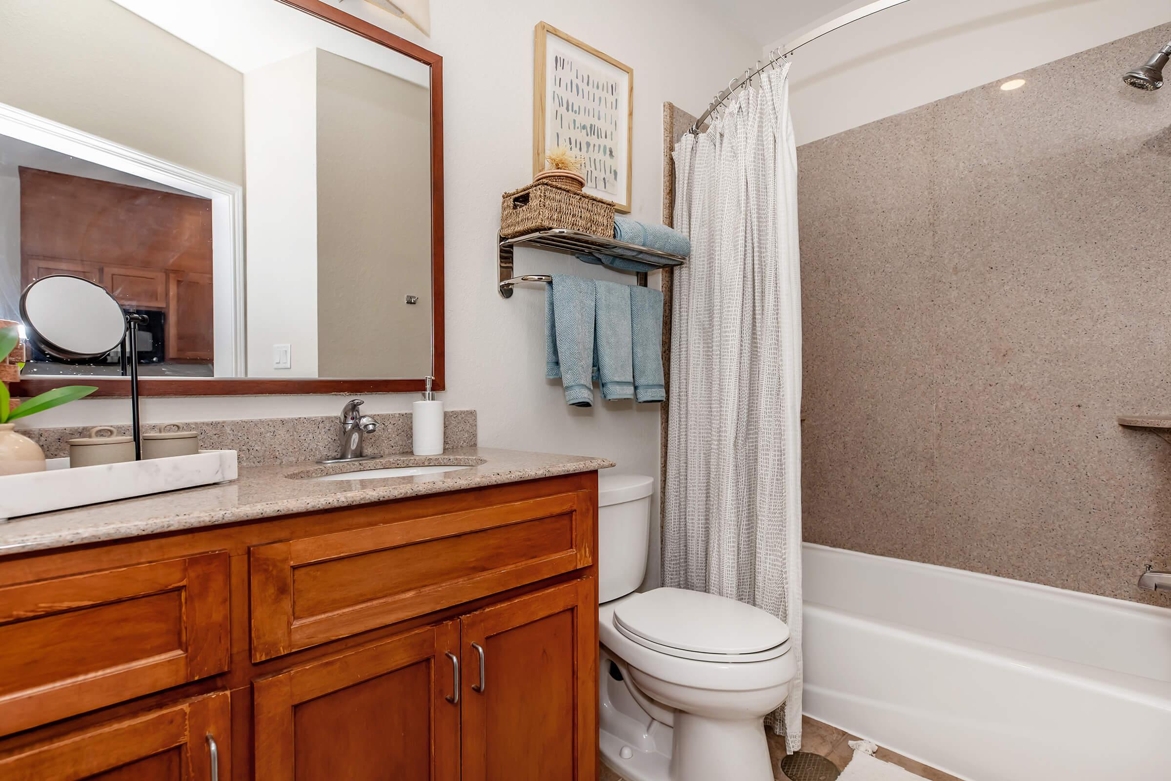 A compact bathroom featuring a wooden vanity with a sink, a mirror above it, and a shower area with a curtain. The decor includes a woven basket and towels hanging on a rack. The walls are painted in neutral tones, and the floor is tiled, creating a clean and modern atmosphere.