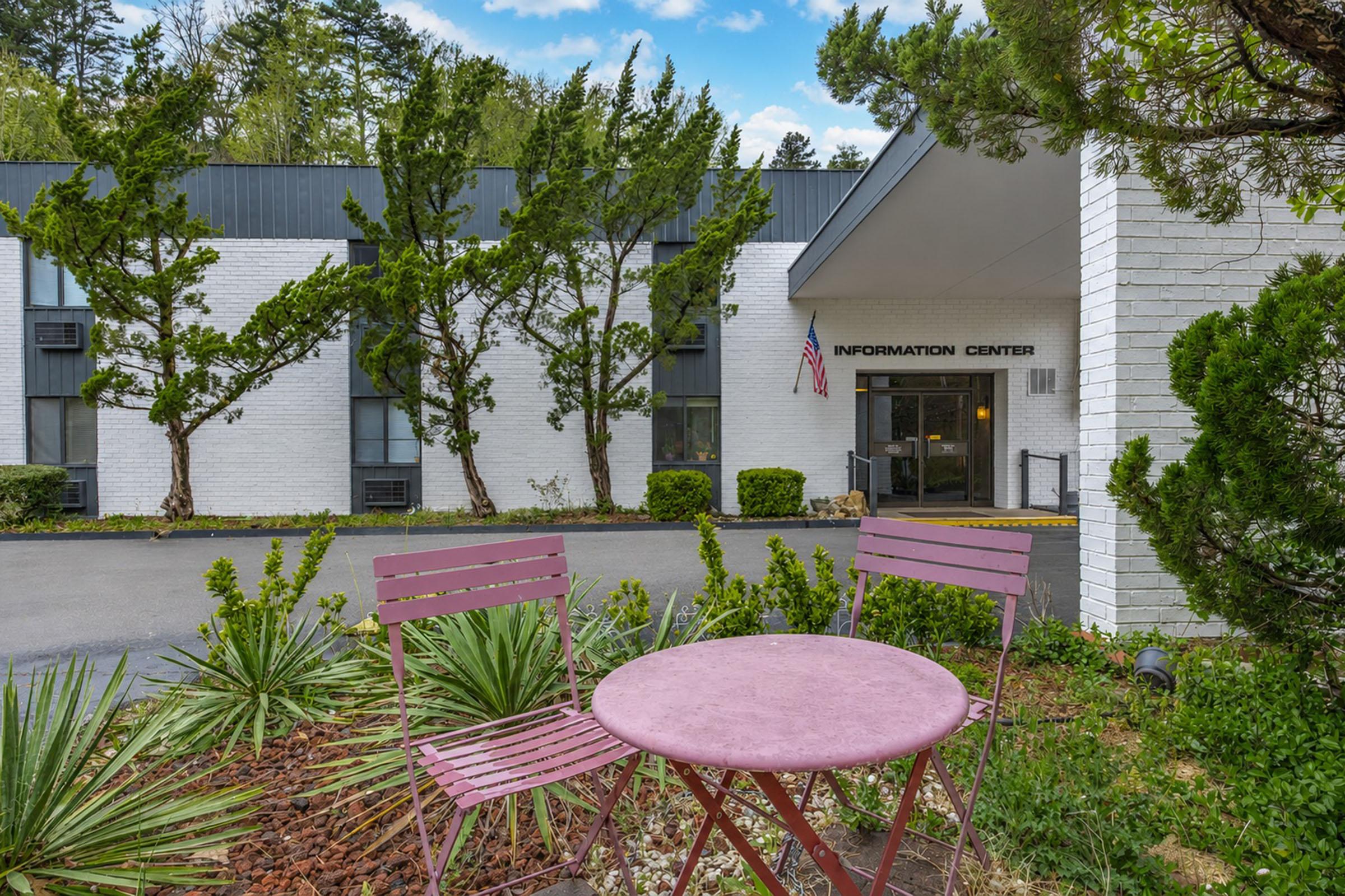Exterior view of a building labeled "Information Center," featuring a welcoming entrance with an American flag. In the foreground, there's a small purple table with two matching chairs, surrounded by landscaped greenery and shrubs. The setting appears inviting and well-maintained, nestled among trees.