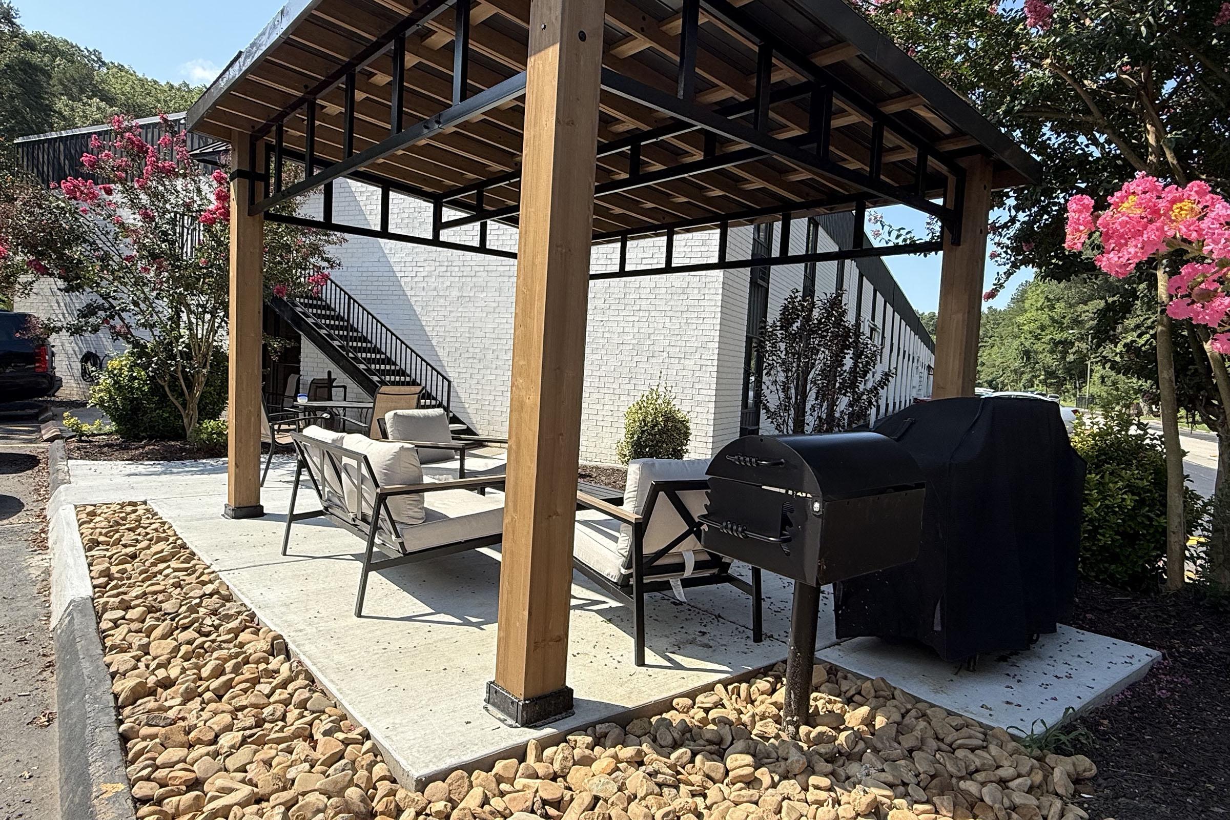 A shaded outdoor seating area featuring modern furniture, a charcoal grill, and landscaping with decorative rocks. The background includes a building and greenery, creating a relaxing environment for gatherings.