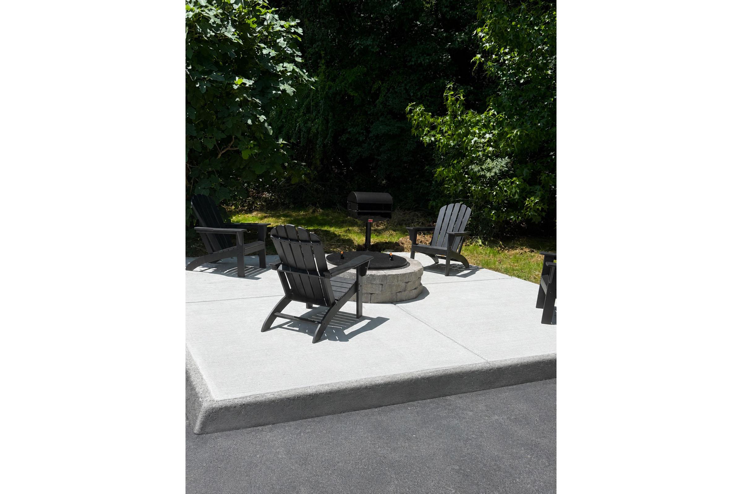 A cozy outdoor fire pit area featuring gray stone seating, surrounded by lush greenery. Four black Adirondack chairs are arranged around the circular fire pit, positioned on a concrete surface. The scene is bright and inviting, perfect for gatherings or relaxing evenings outdoors.