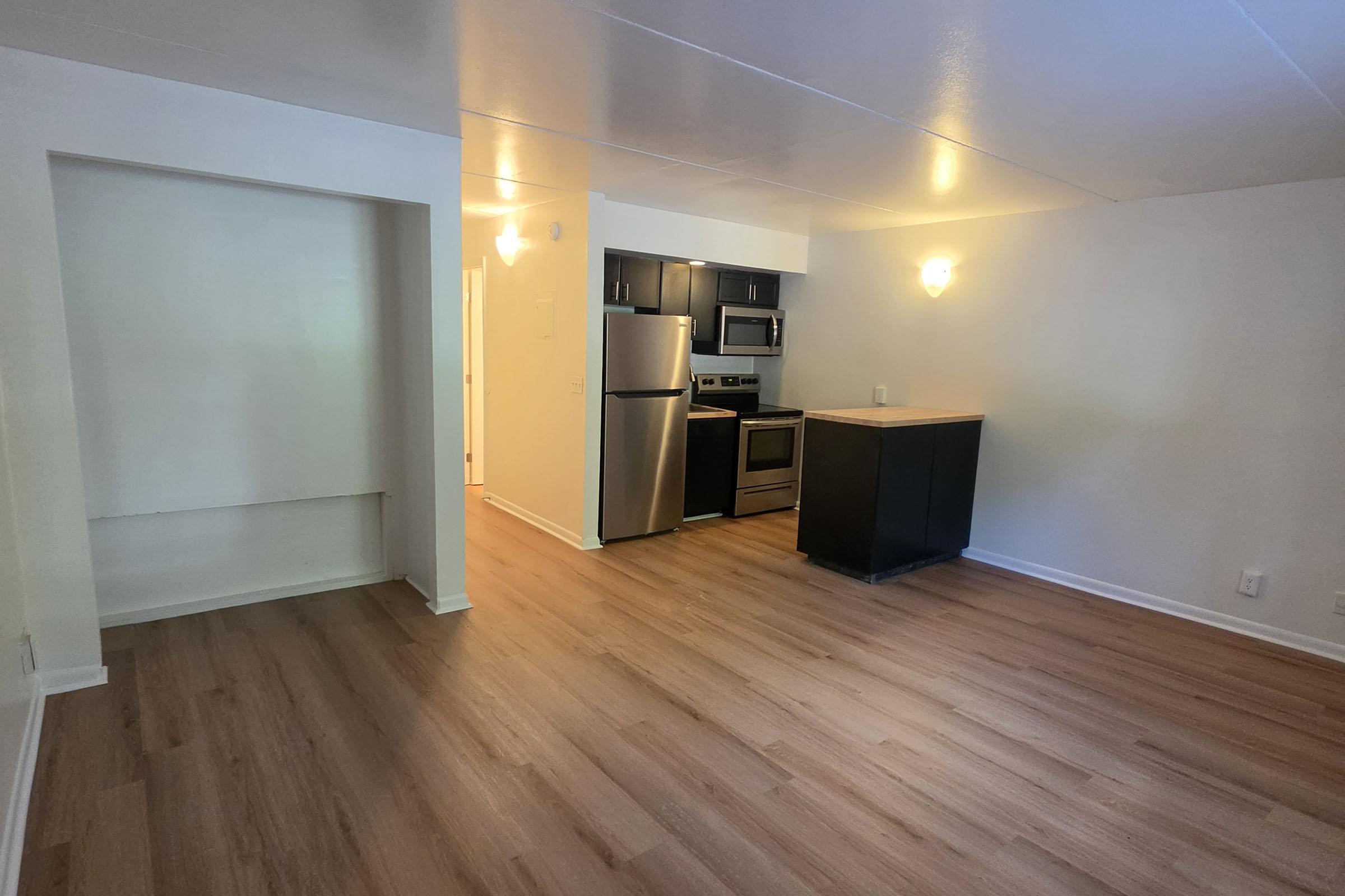 A modern kitchen and living area with laminate flooring, featuring a black kitchen island, stainless steel appliances, and light-colored walls. The space is well-lit with wall sconces, and an alcove is visible to the left. The overall design is open and inviting, suitable for a contemporary living space.
