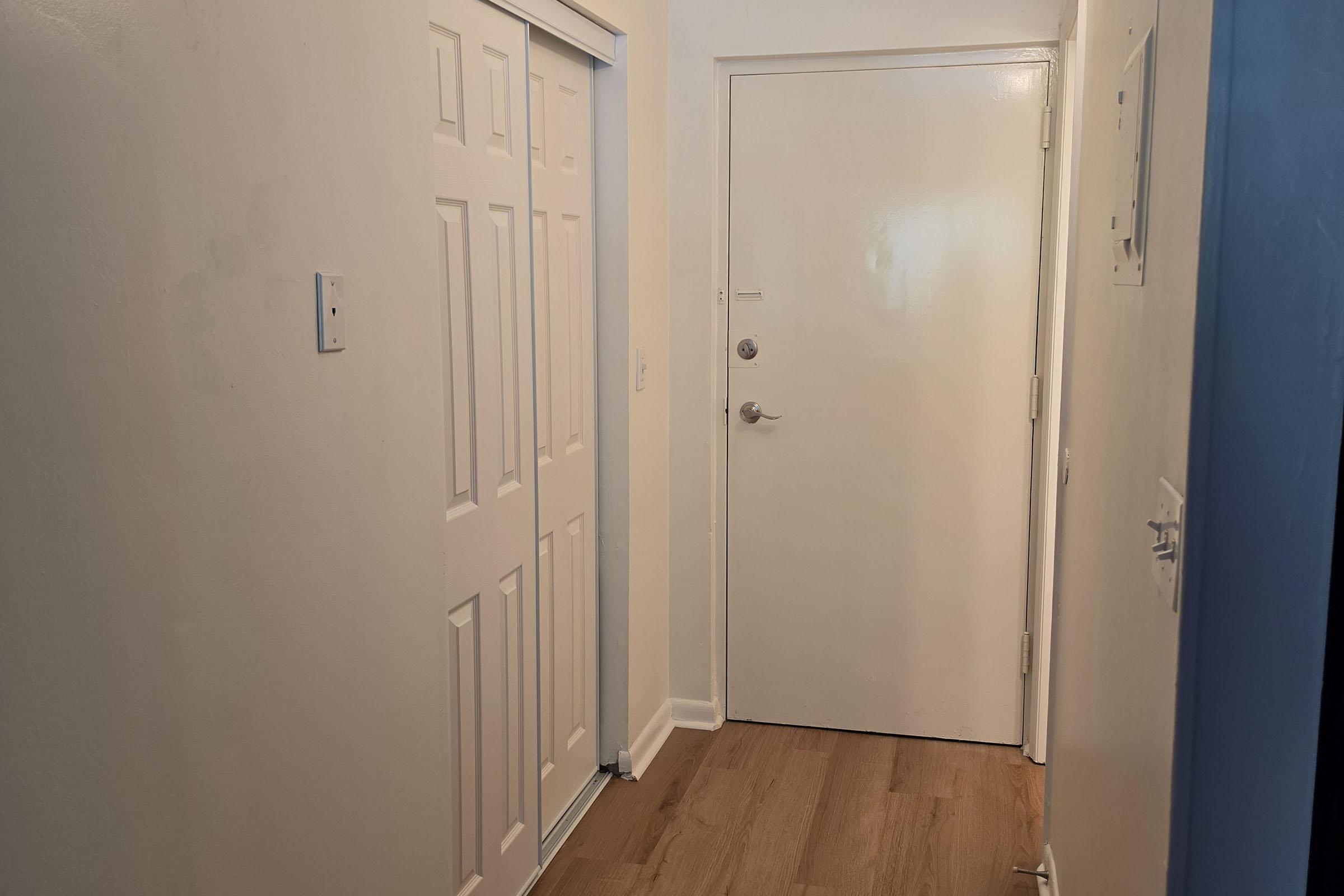 A well-lit hallway featuring a closed white door on the left, a white wall, and a light wood floor. The main door is visible on the right, with a simple handle and no visible decorations. The walls are painted white, creating a clean and modern look.