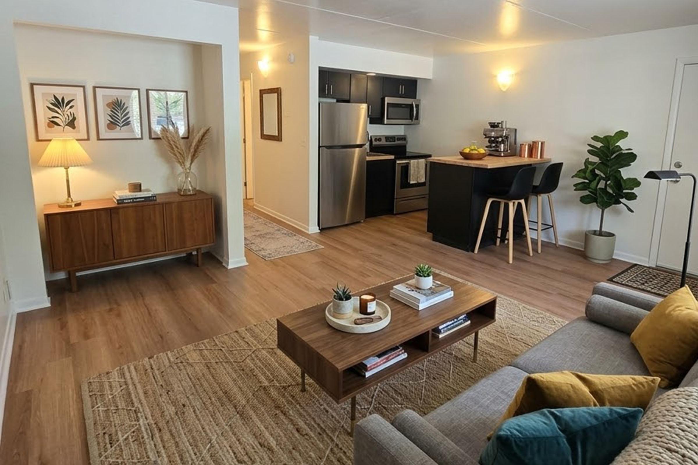 A bright, modern living space featuring an open kitchen and living area. The room has wooden flooring, a cozy gray sofa with yellow and green accent pillows, a wooden coffee table with decorations, a stylish lamp, and indoor plants. The kitchen is equipped with stainless steel appliances and a bar with two stools.
