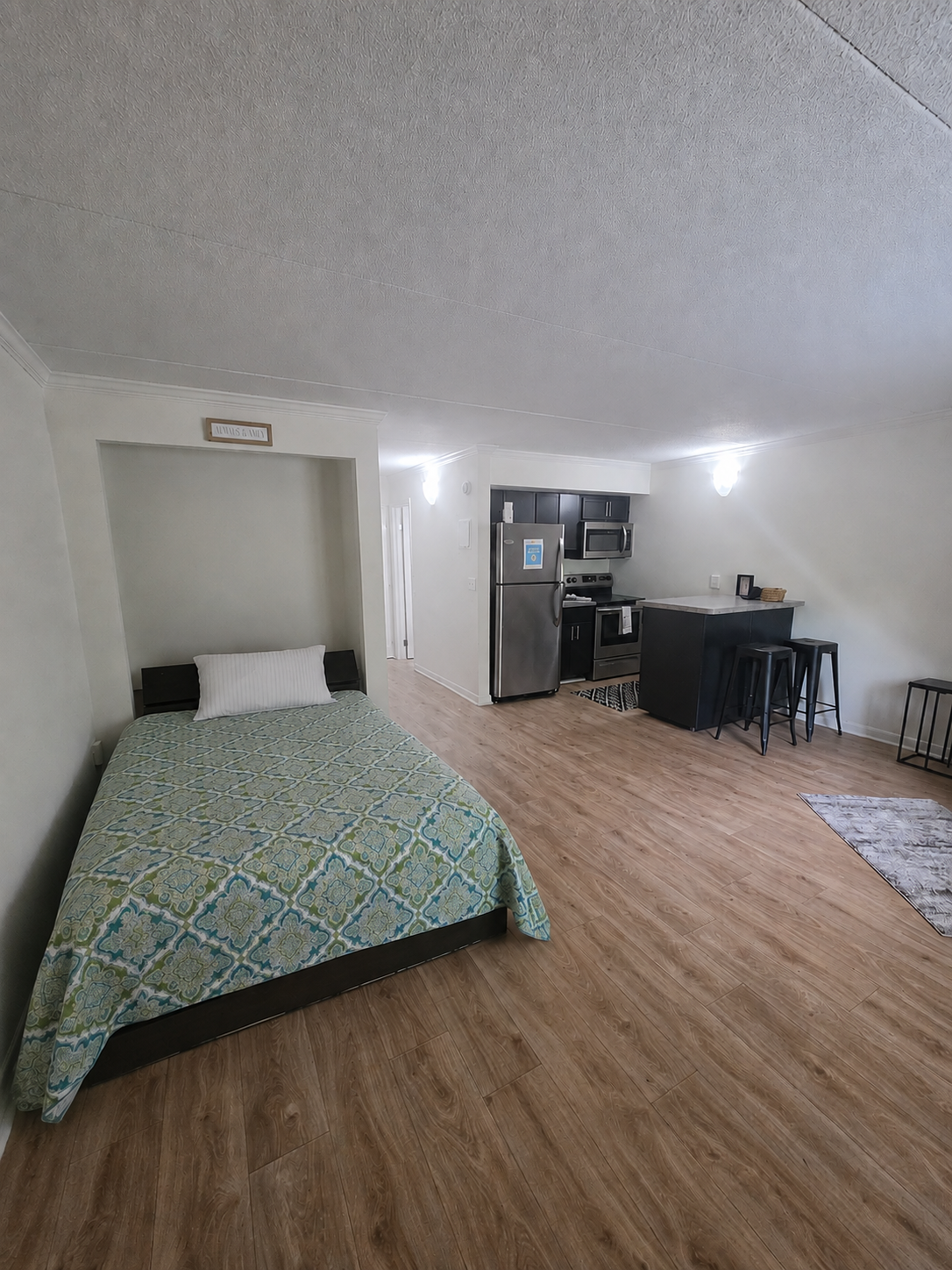 A modern, minimalist studio apartment featuring a bed with a colorful patterned blanket, a small kitchen area with stainless steel appliances, a bar with stools, and a light-colored wooden floor. The walls are painted in soft tones, and the space is well-lit with natural and overhead lighting.