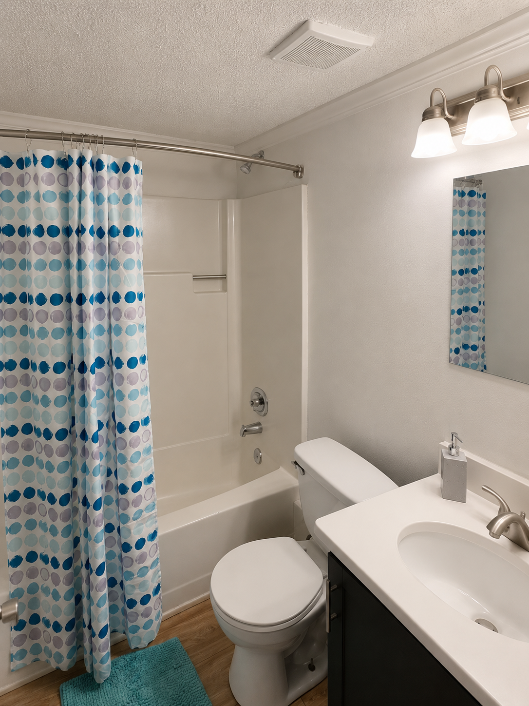 A clean bathroom featuring a shower and tub combination, a white toilet, and a modern sink with a countertop. The shower curtain has a blue and turquoise polka dot pattern, and there is a small blue rug on the floor. The lighting is bright, highlighting the simple, fresh design.